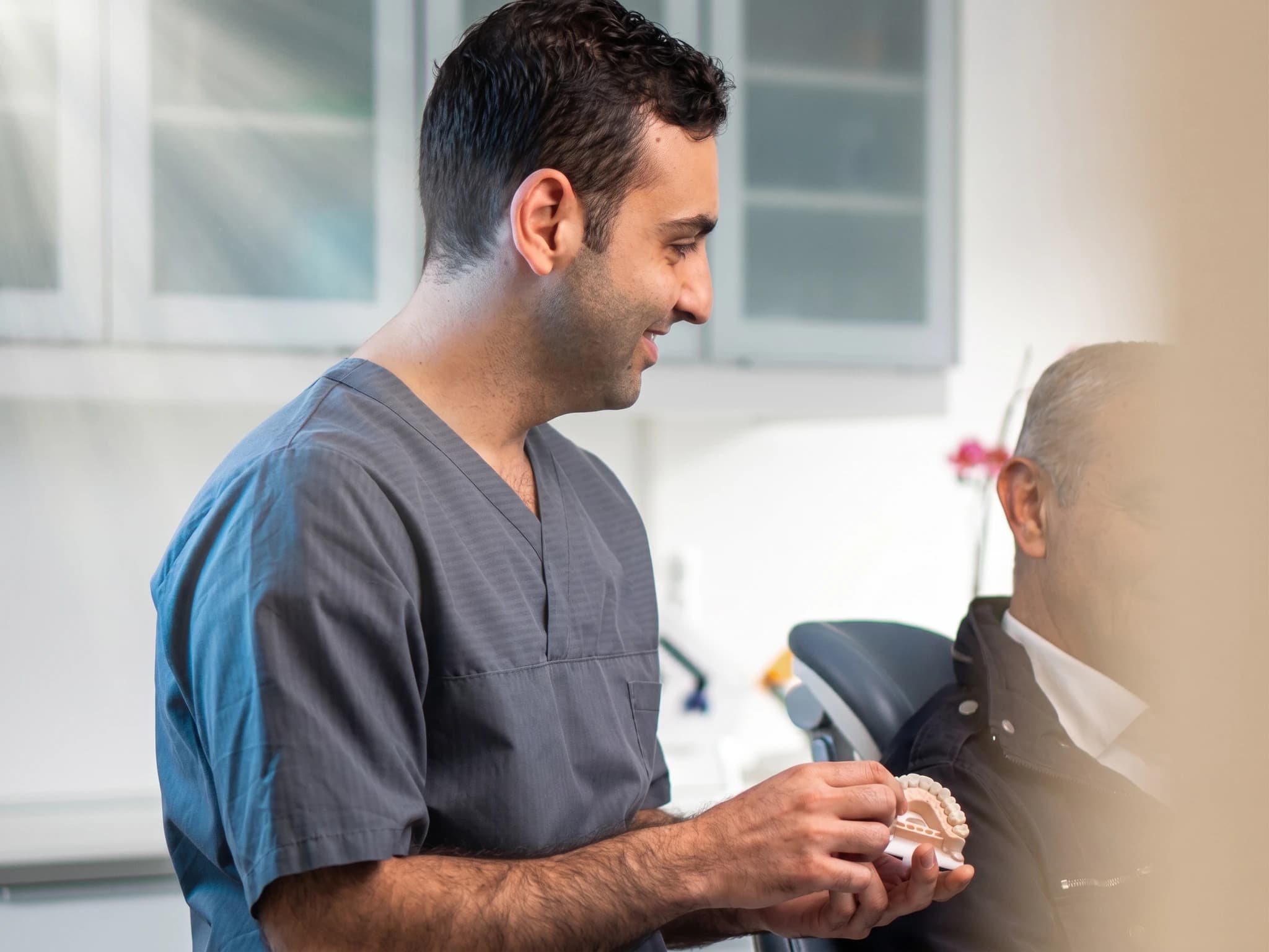 Tandläkare i grå scrubs förklarar en tandmodell för en patient som sitter i en tandläkarstol.