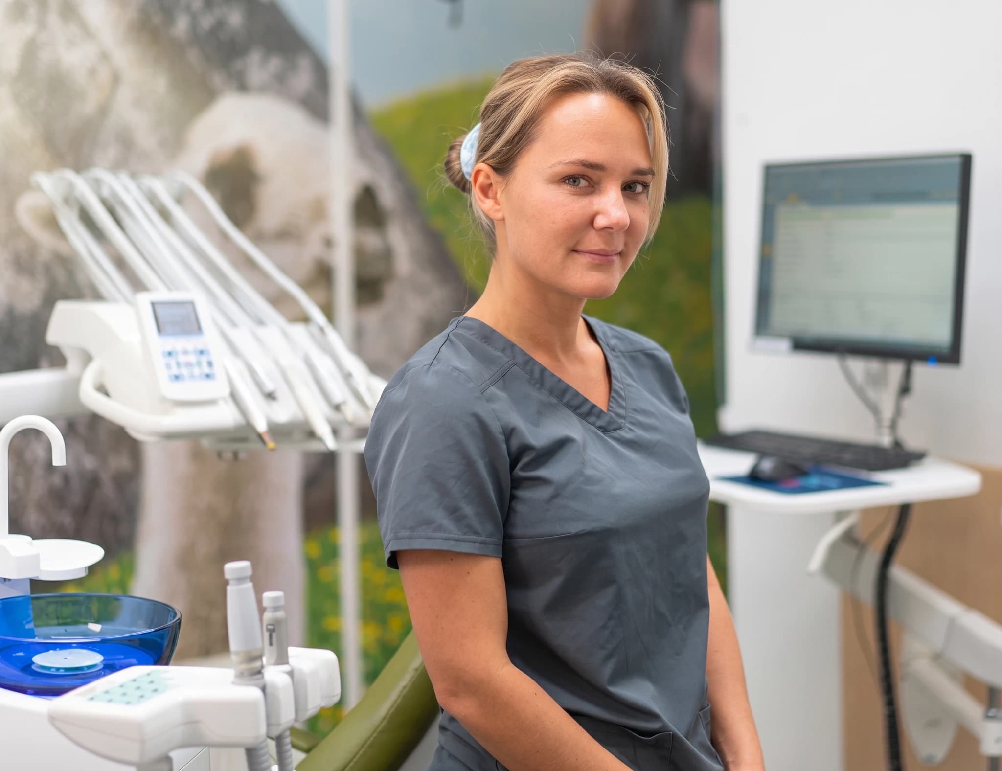 Tandläkare i grå scrubs ler mot kameran i en modern klinik, med tandvårdsutrustning och en datorskärm i bakgrunden.