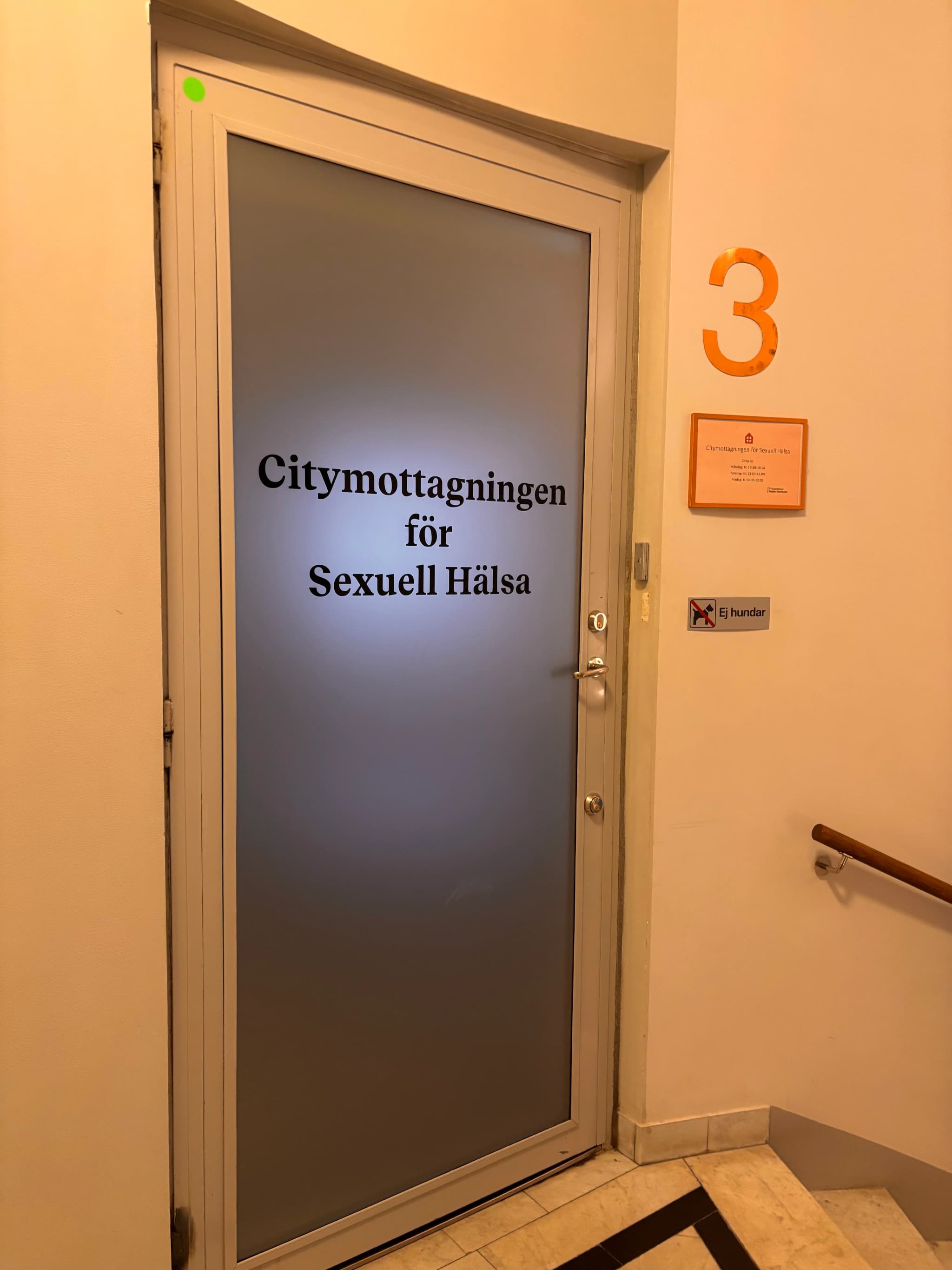En frostad glasdörr med texten "Citymottagningen för Sexuell Hälsa" på svenska, samt skyltar med öppettider och information om att hundar inte är tillåtna.