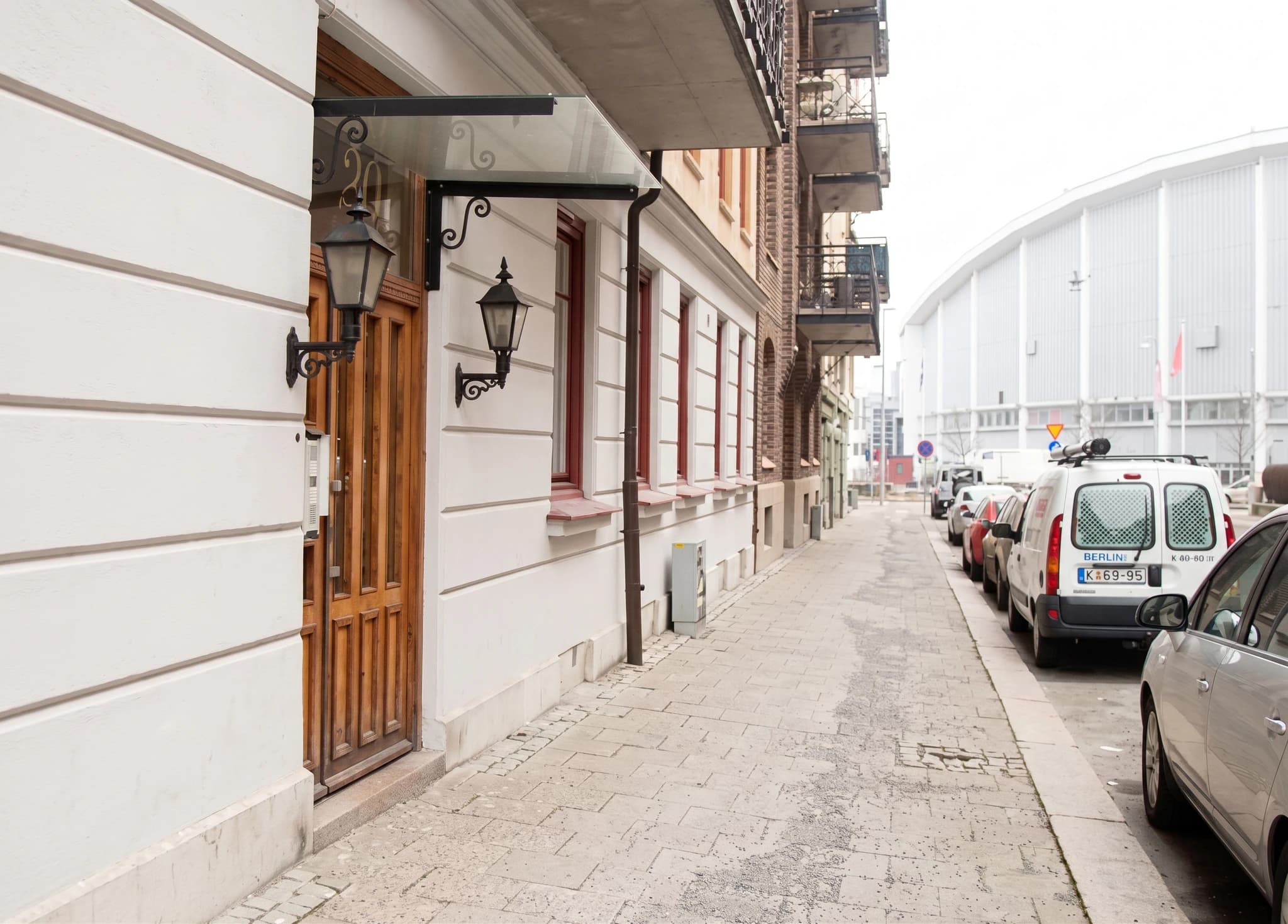 Entrébyggnad med trädörr och fönster, samt en trottoar med parkerade bilar och en modern byggnad i bakgrunden.