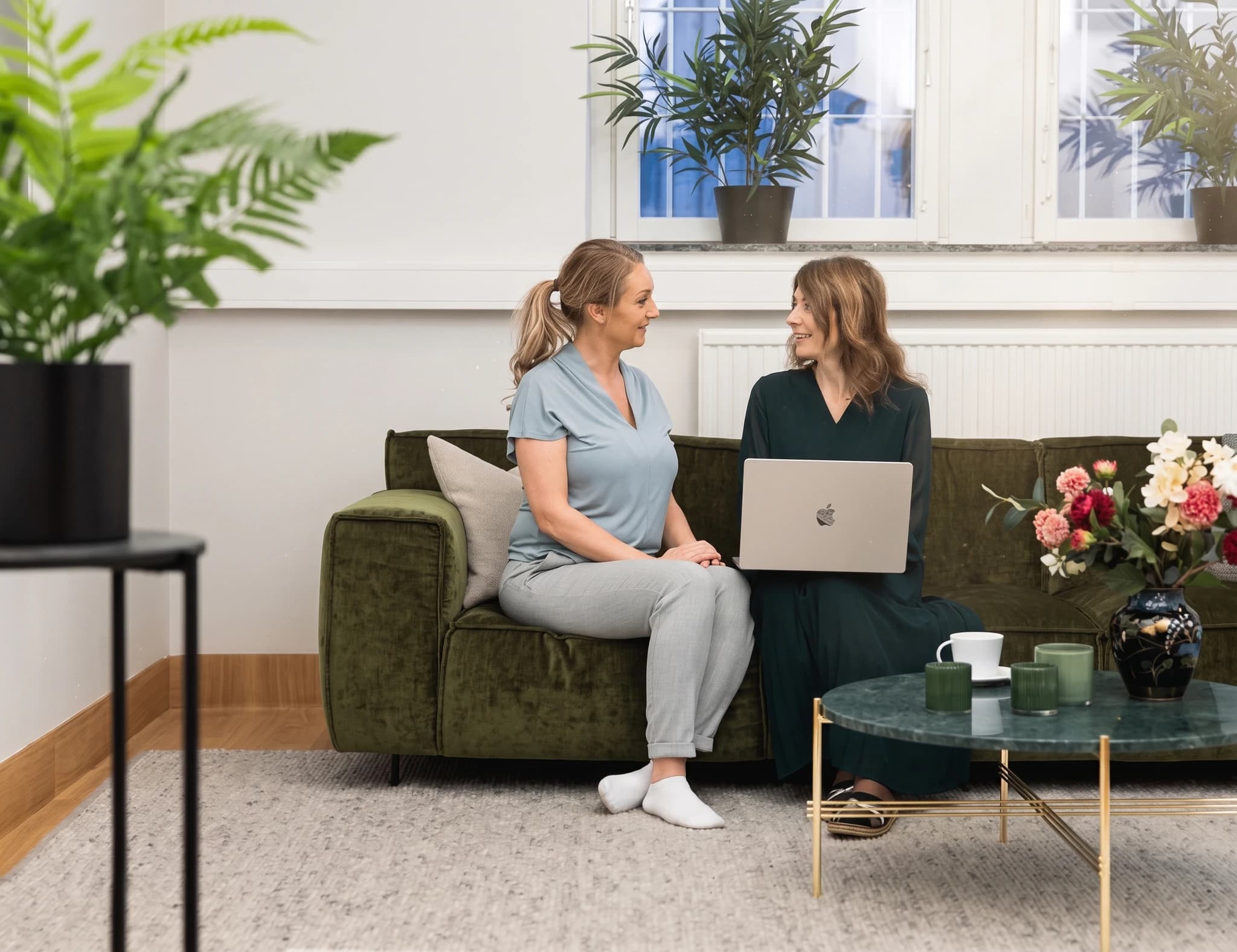 Två kvinnor sitter på en grön soffa och pratar medan en av dem håller en laptop.