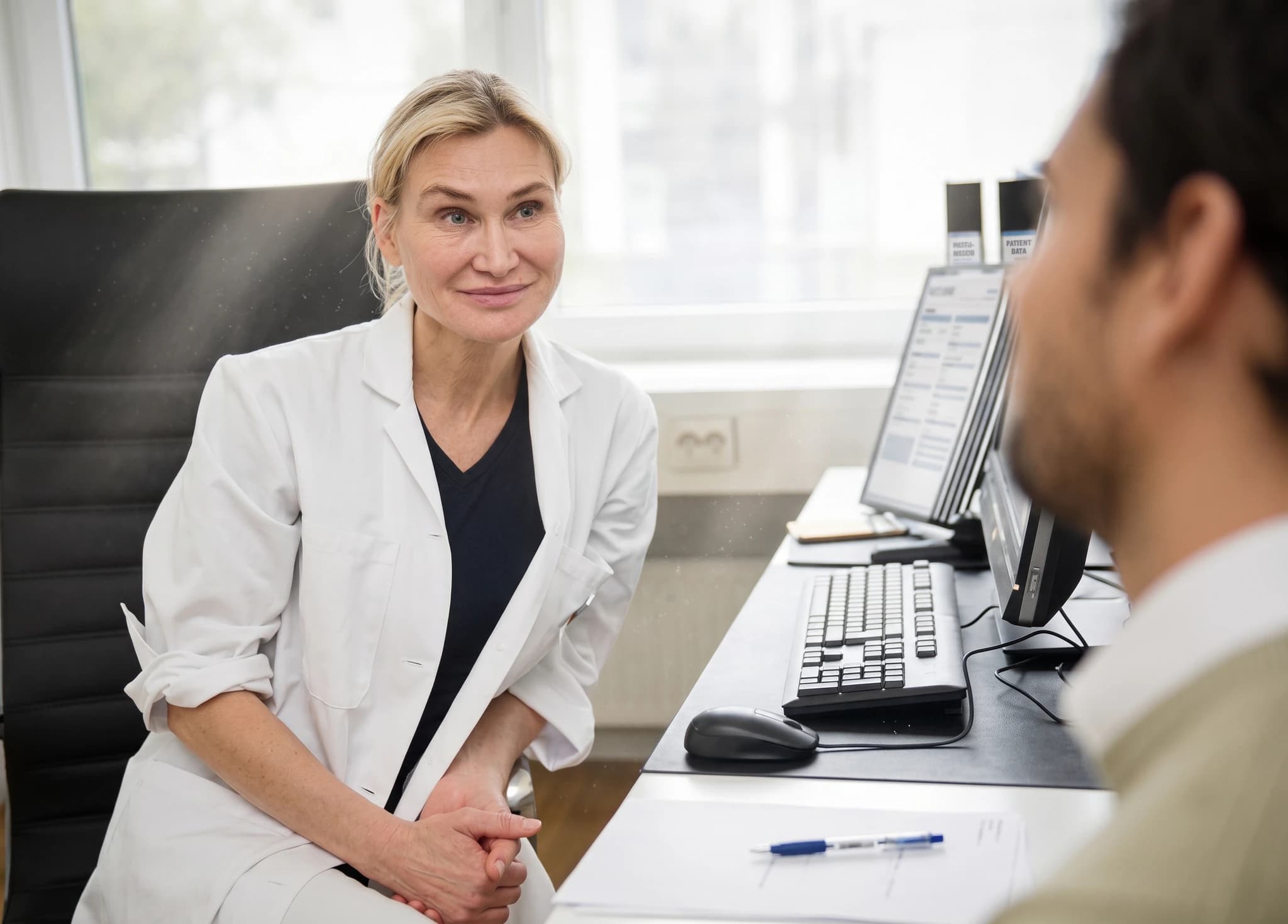 En kvinnlig läkare i vit rock sitter och pratar med en patient på ett kontor.