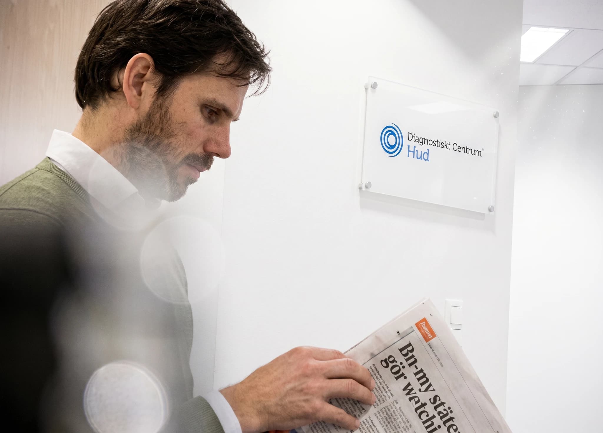 Man med skägg i kostym och kavaj står och läser en tidning framför en vägg med en skylt för Diagnostiskt Centrum Hud.