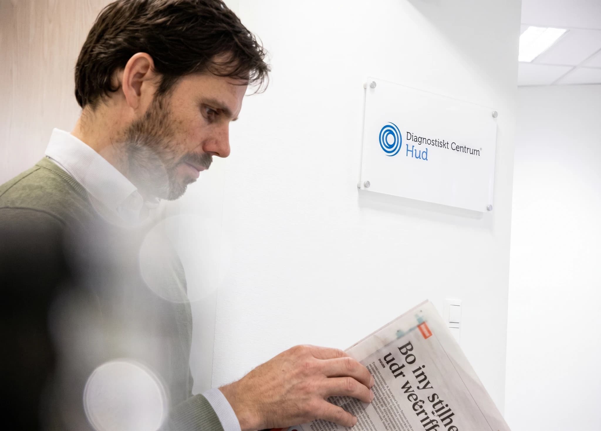 Man med skägg läser en tidning i en klinikhall, med en skylt för 'Diagnostiskt Centrum Hud' på väggen.