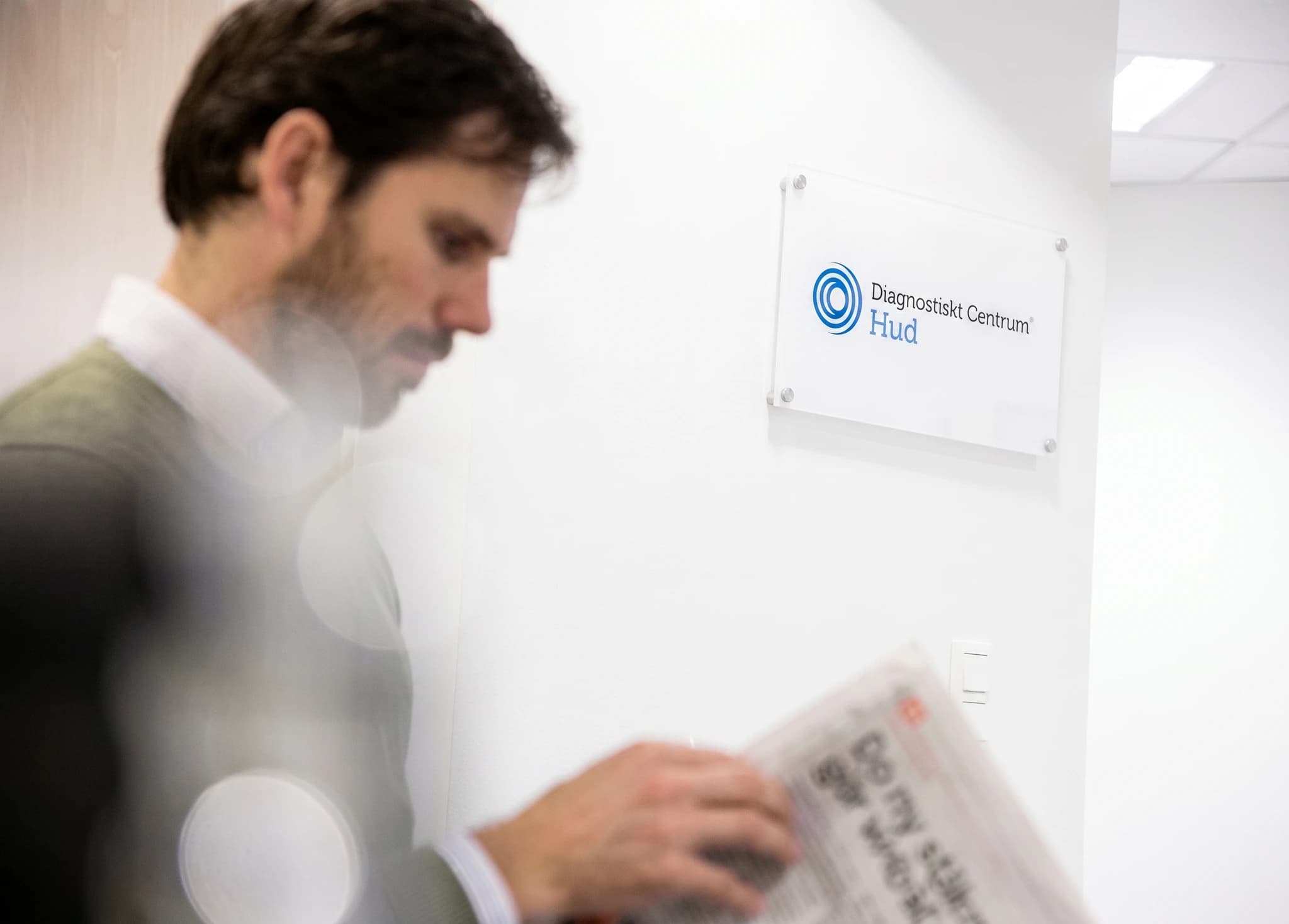 Man i kavaj läser tidning i en ljus korridor framför en skylt för Diagnostiskt Centrum Hud.