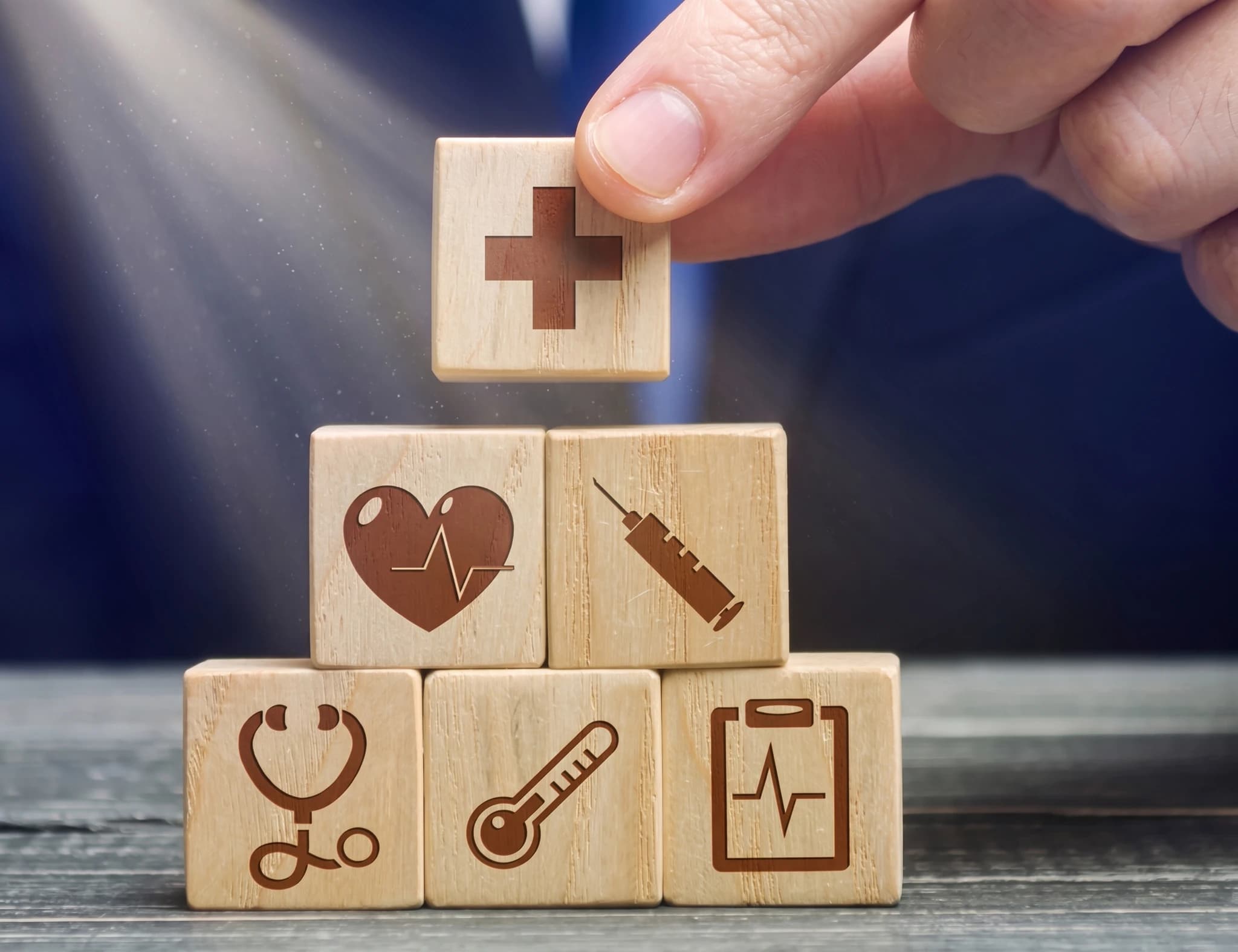 En hand placerar en träkloss med ett rött korssymbol ovanpå en pyramid av sex träklossar med hälsorelaterade symboler: ett hjärta med EKG-linje, en spruta, ett stetoskop, en termometer och ett clipboard.
