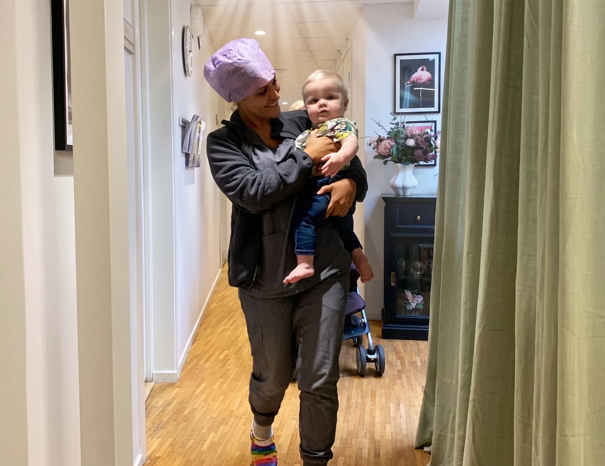 Sjuksköterska med lila mössa bär ett litet barn i famnen medan hon går i en korridor på en klinik.