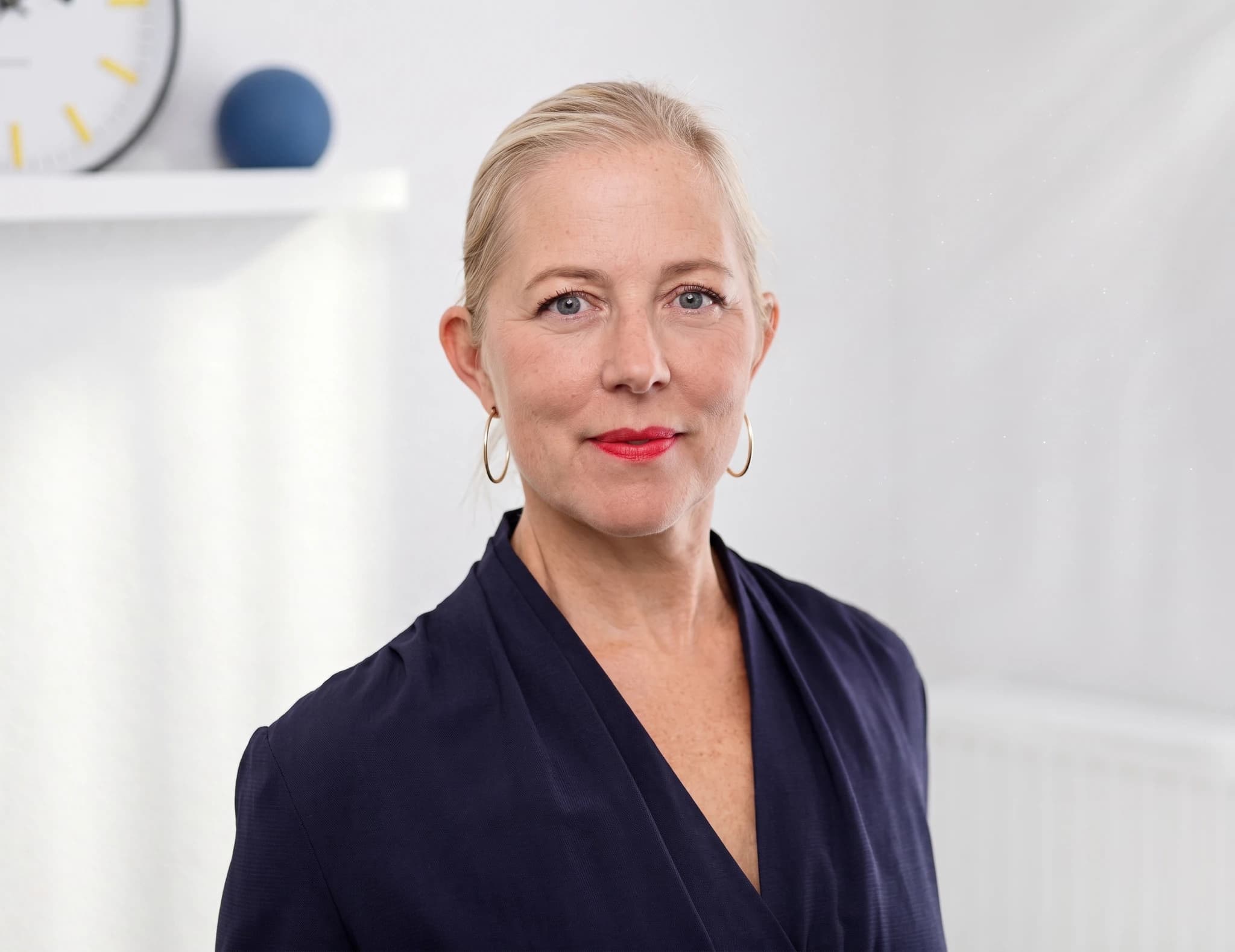 Porträtt av en blond kvinna med hoopörhängen och en mörkblå blus, med en vit kontorsbakgrund.