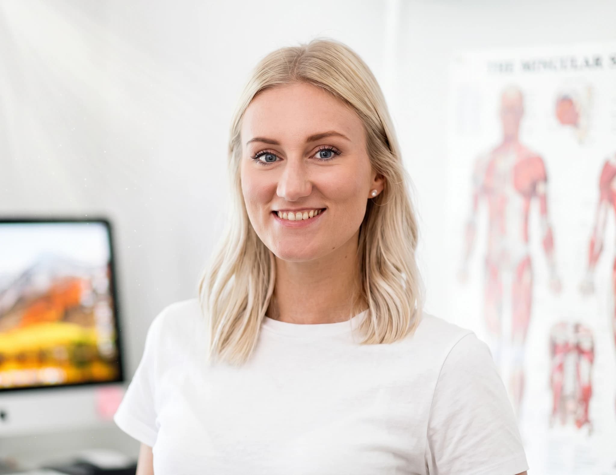 En kvinna med blont hår ler mot kameran i ett kontor, med en datorskärm och en anatomisk affisch i bakgrunden.