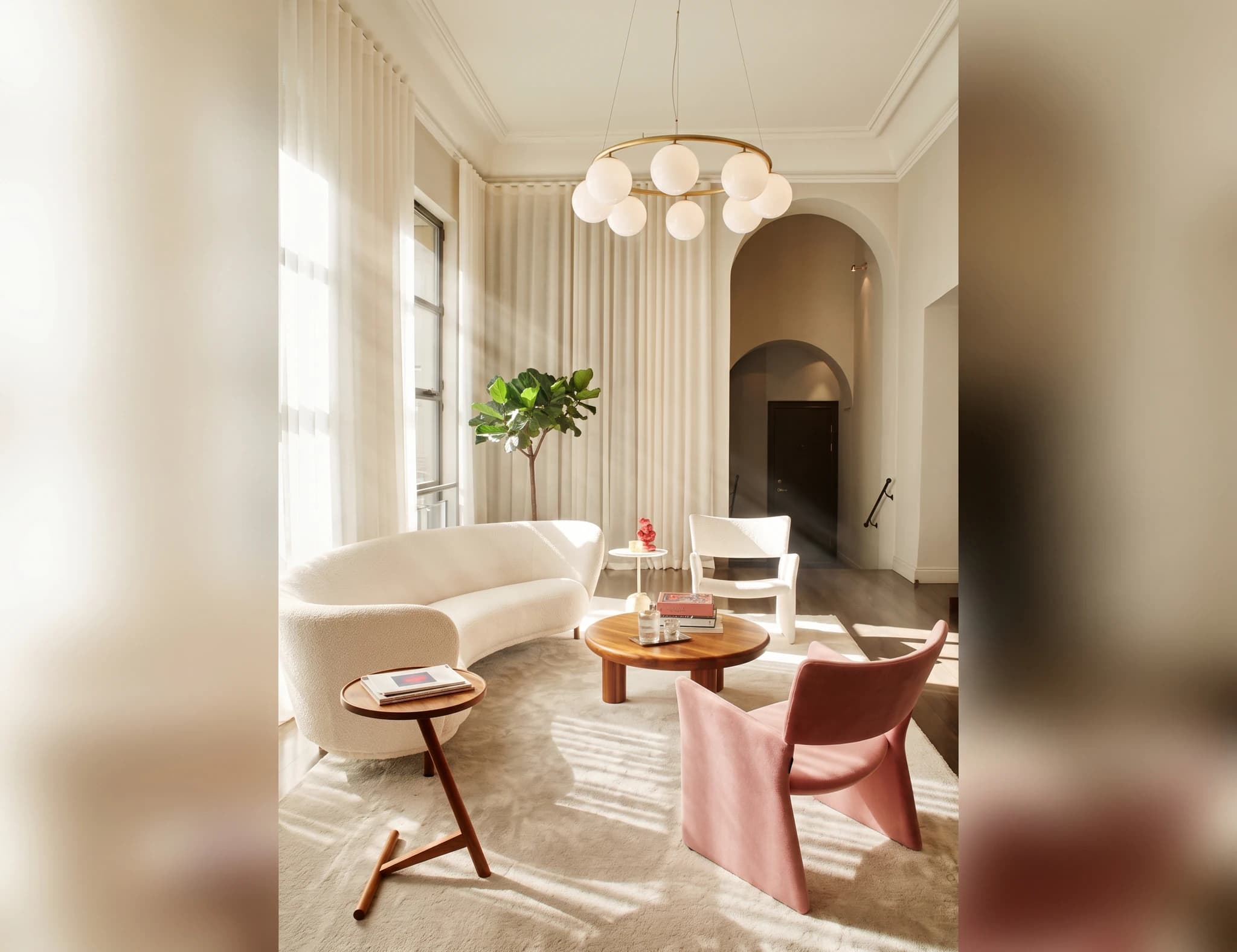 Modernt vardagsrum med en krökt vit soffa, en rosa fåtölj, ett runt soffbord och stora vita draperier som släpper in solljus.