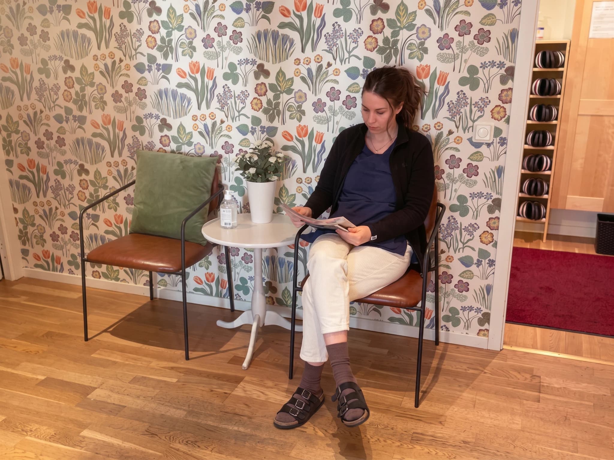 En kvinna sitter och läser en tidning i ett väntrum med blommig tapet.