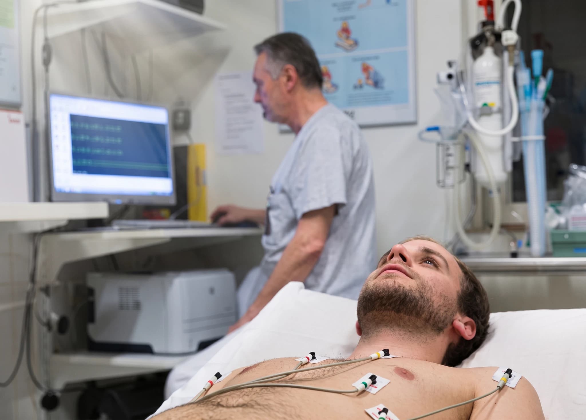 Patient med EKG-elektroder på bröstet ligger i sängen medan en läkare sitter vid en dator i bakgrunden.