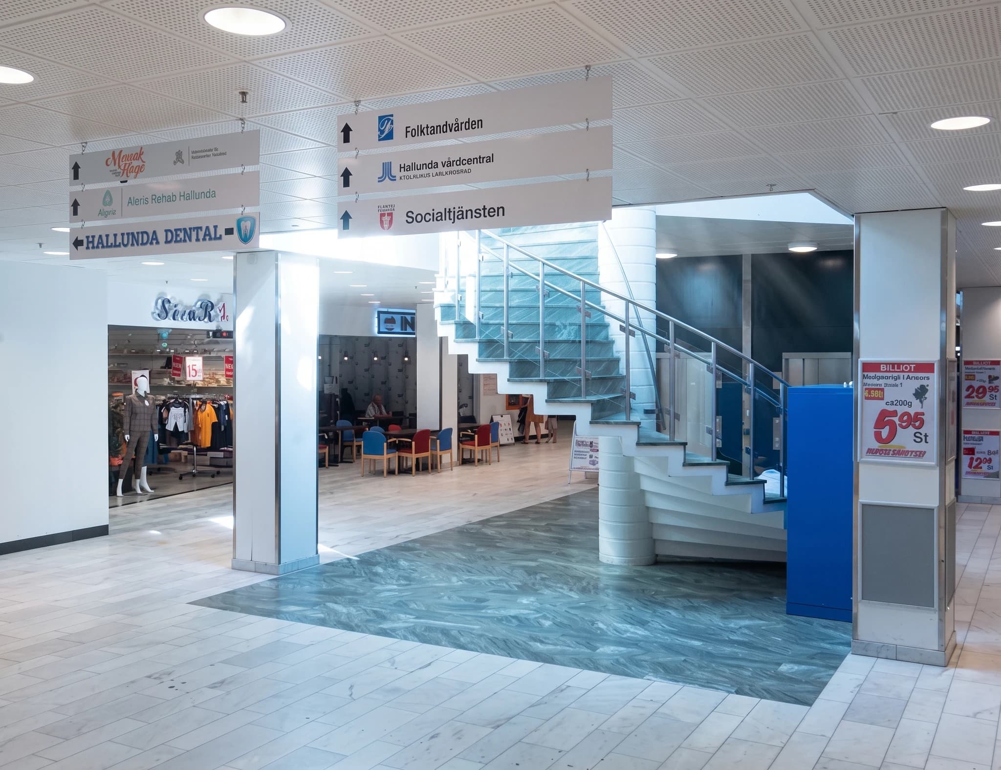 En interiörbild av en inomhuskorridor med flera skyltar som pekar ut olika avdelningar på en vårdcentral, inklusive "Folktandvården", "Hallunda vårdcentral" och "Socialtjänsten".