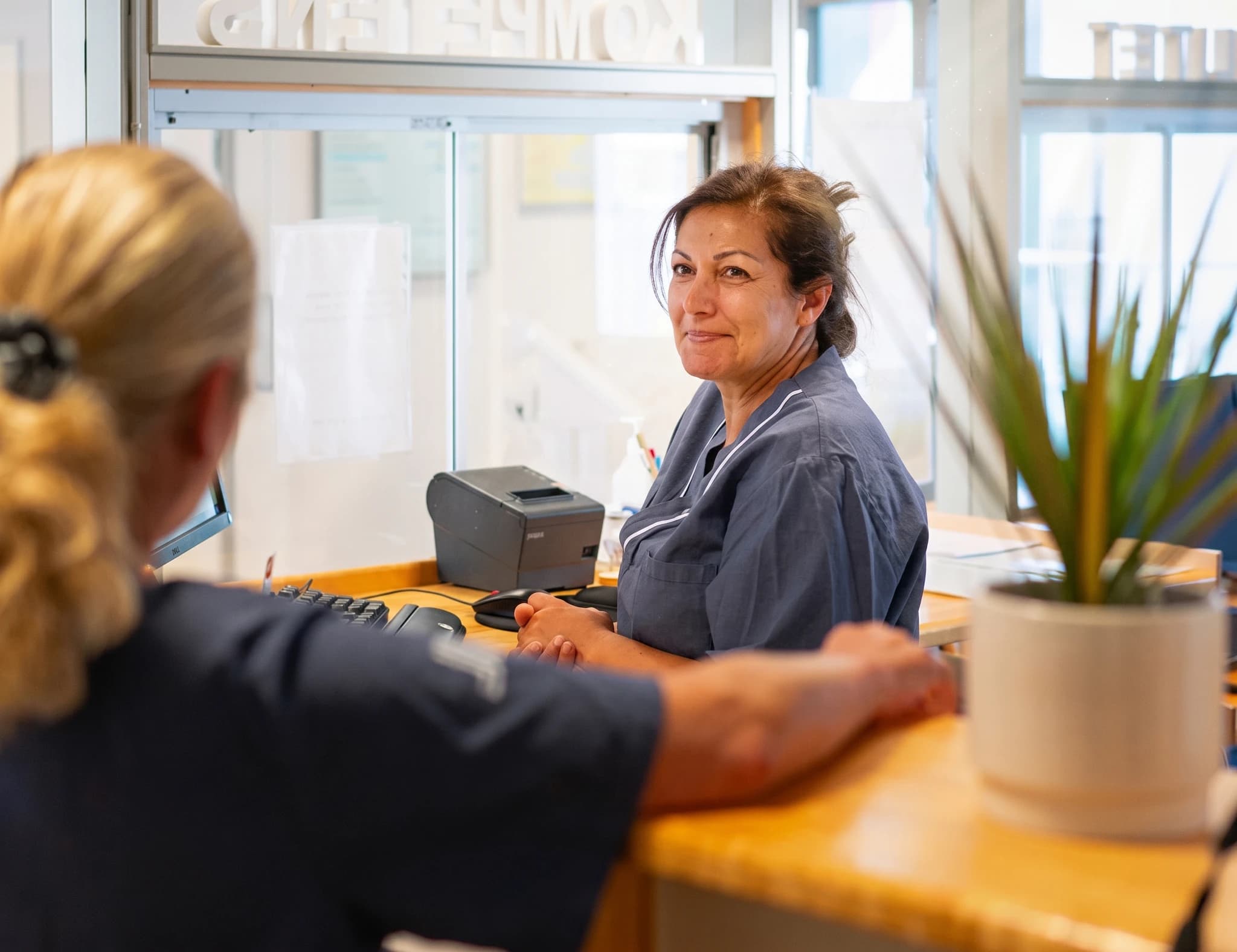 En receptionist med mörkbrunt hår ler medan hon vänder sig mot kameran bakom en reception, där en person till höger sitter vid en datorskärm och tangentbord.