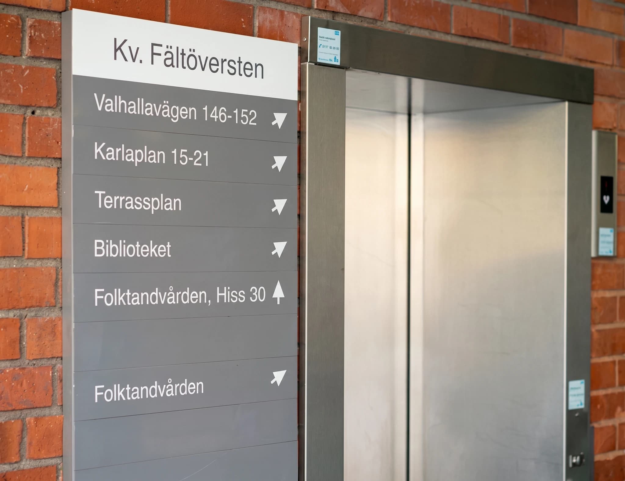 En grå informationsskylt med en lista över destinationer och pappersflygplan som indikerar riktningen, placerad bredvid en hiss i en tegelbyggnad.