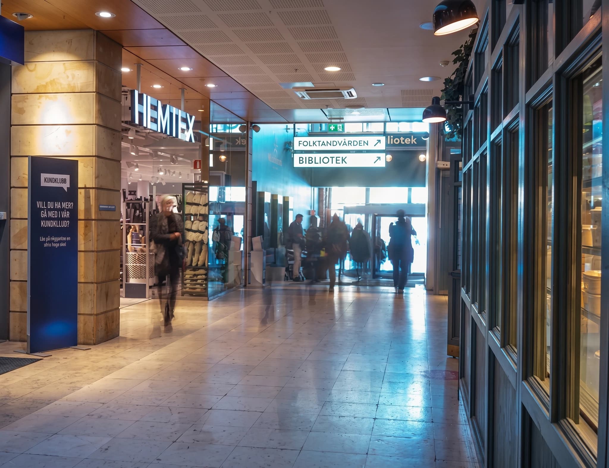 Människor går genom en inomhus galleria med skyltar för folktandvården och biblioteket.