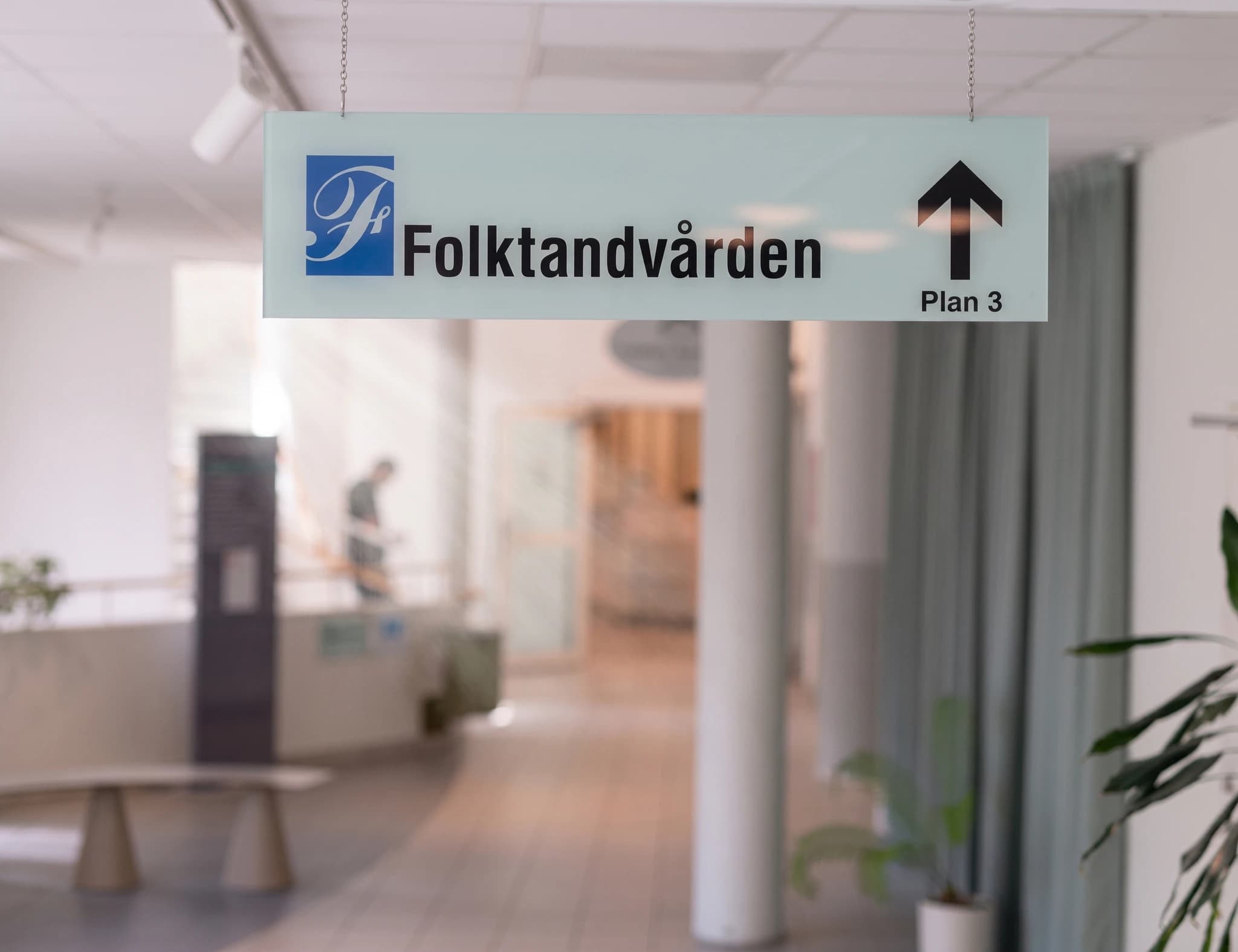 En skylt med texten "Folktandvården" och en pil som pekar uppåt mot "Plan 3" hänger i en ljus korridor på en klinik.