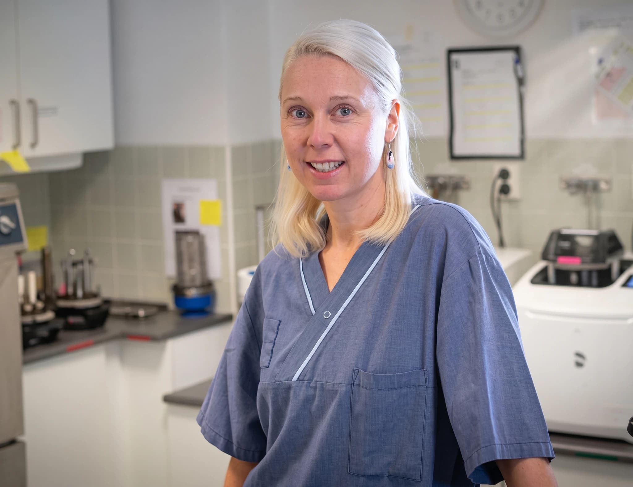 En blond kvinna i blå läkarrock ler mot kameran i ett laboratorium med medicinsk utrustning i bakgrunden.