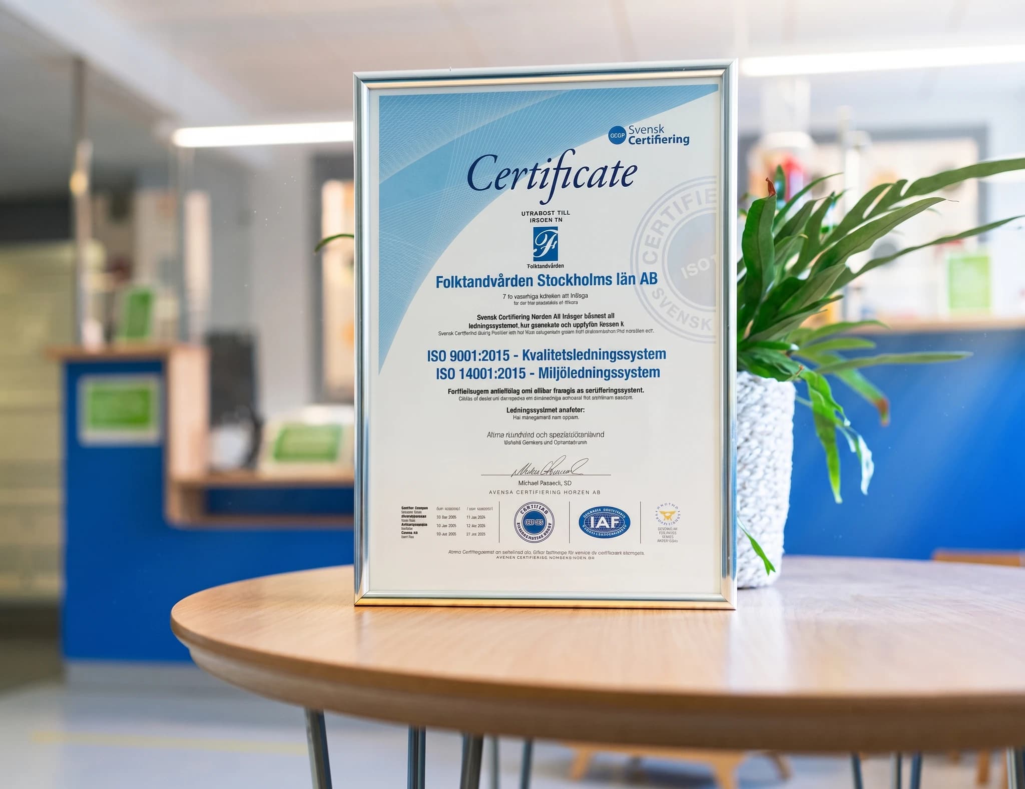 Ett certifikat för ISO 9001:2015 och ISO 14001:2015 från Svensk Certifiering ligger på ett runt träbord framför en växt.