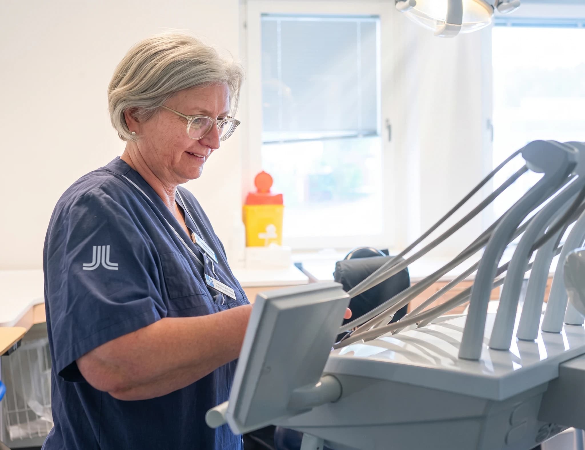 En kvinna i blå sjukvårdskläder med glasögon som arbetar vid kontrollpanelen på en tandvårdsstol.