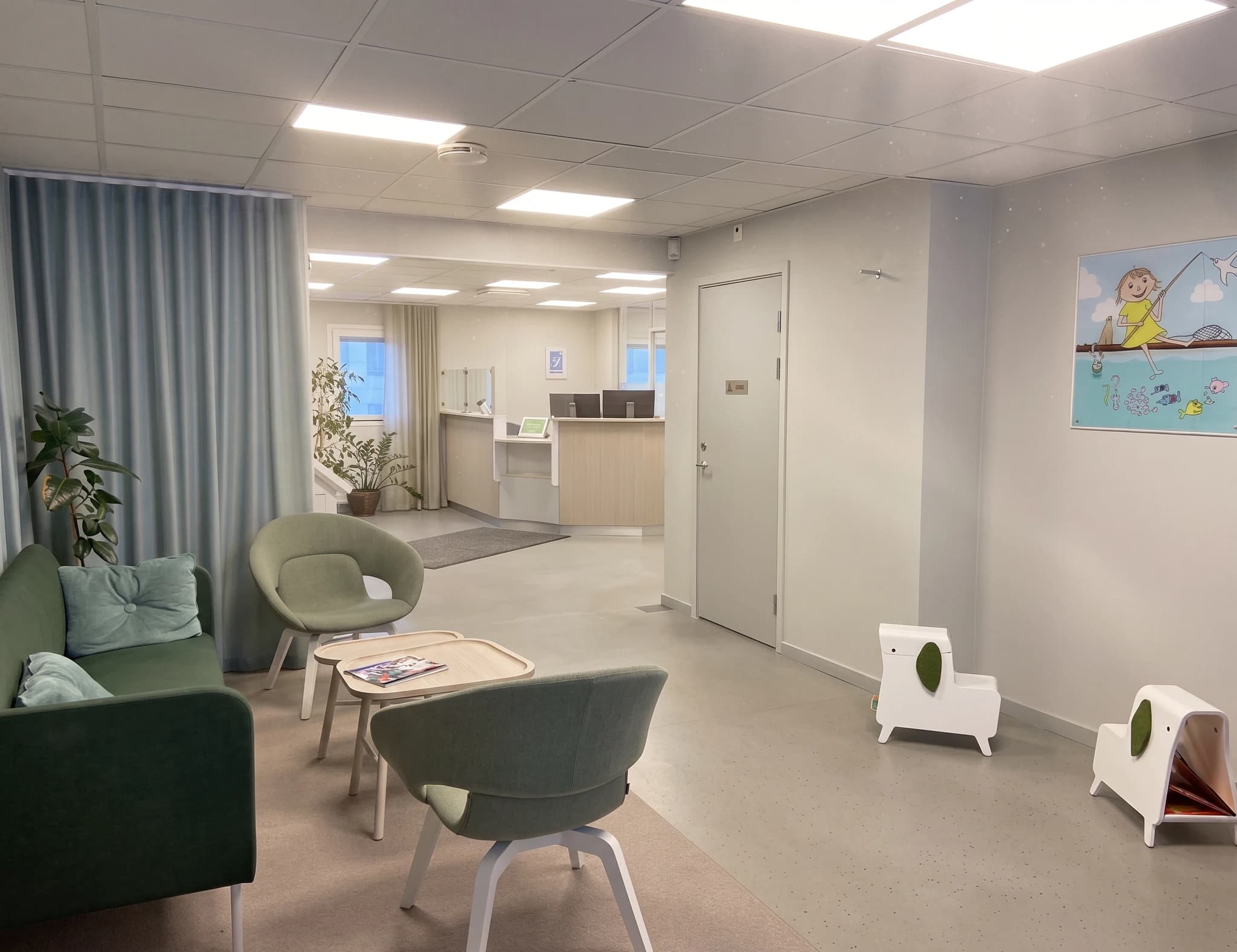 Väntrum med soffor, fåtöljer, låga bord och speciellt sittutrymme för barn, med en receptionsdisk i bakgrunden.