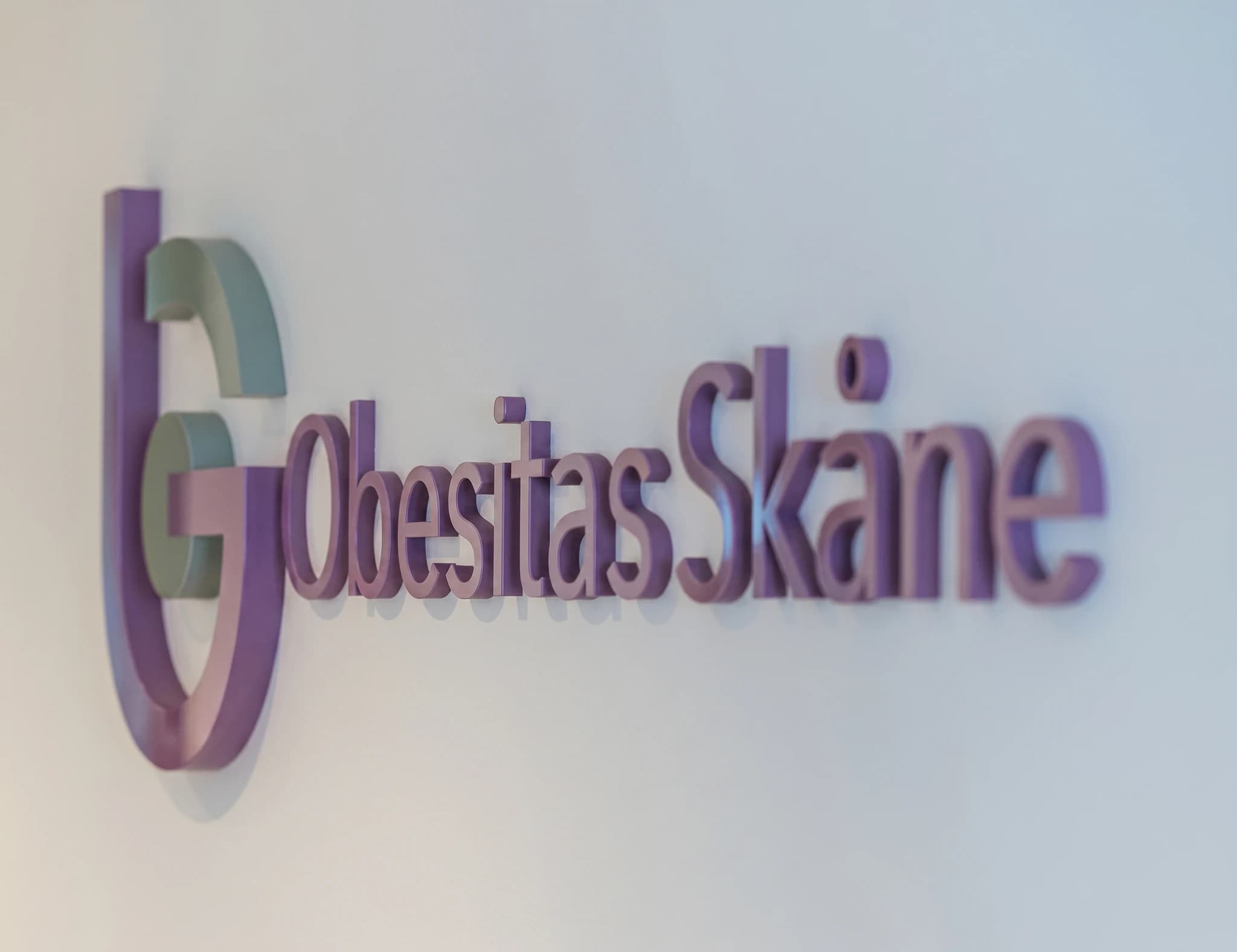 Logotyp för "Obesity Skåne" i lila och grönt på en vit vägg.