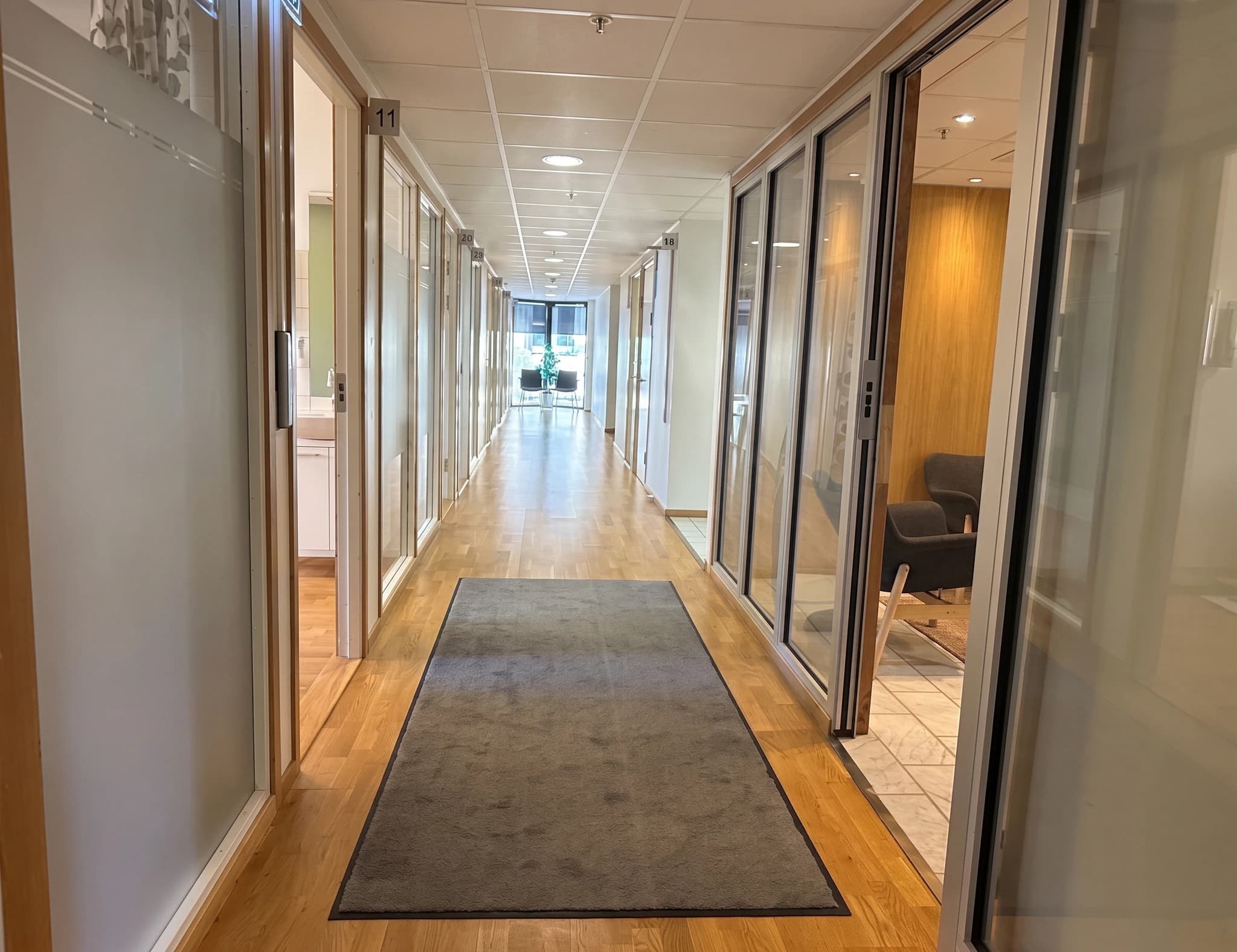 En tom kontorskorridor med glasdörrar till flera mottagningsrum och ett grått gångmatt