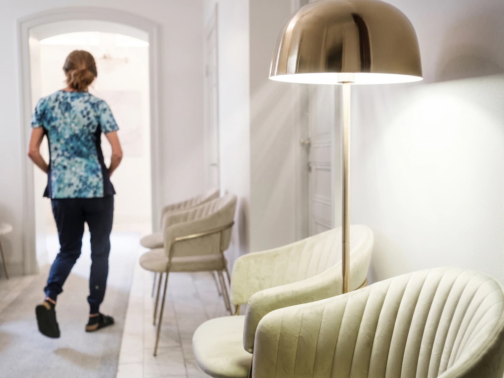 En hälsovårdspersonal i blå och vit mönstrad topp och mörkblå byxor går in i en väntrum och in i en korridor.