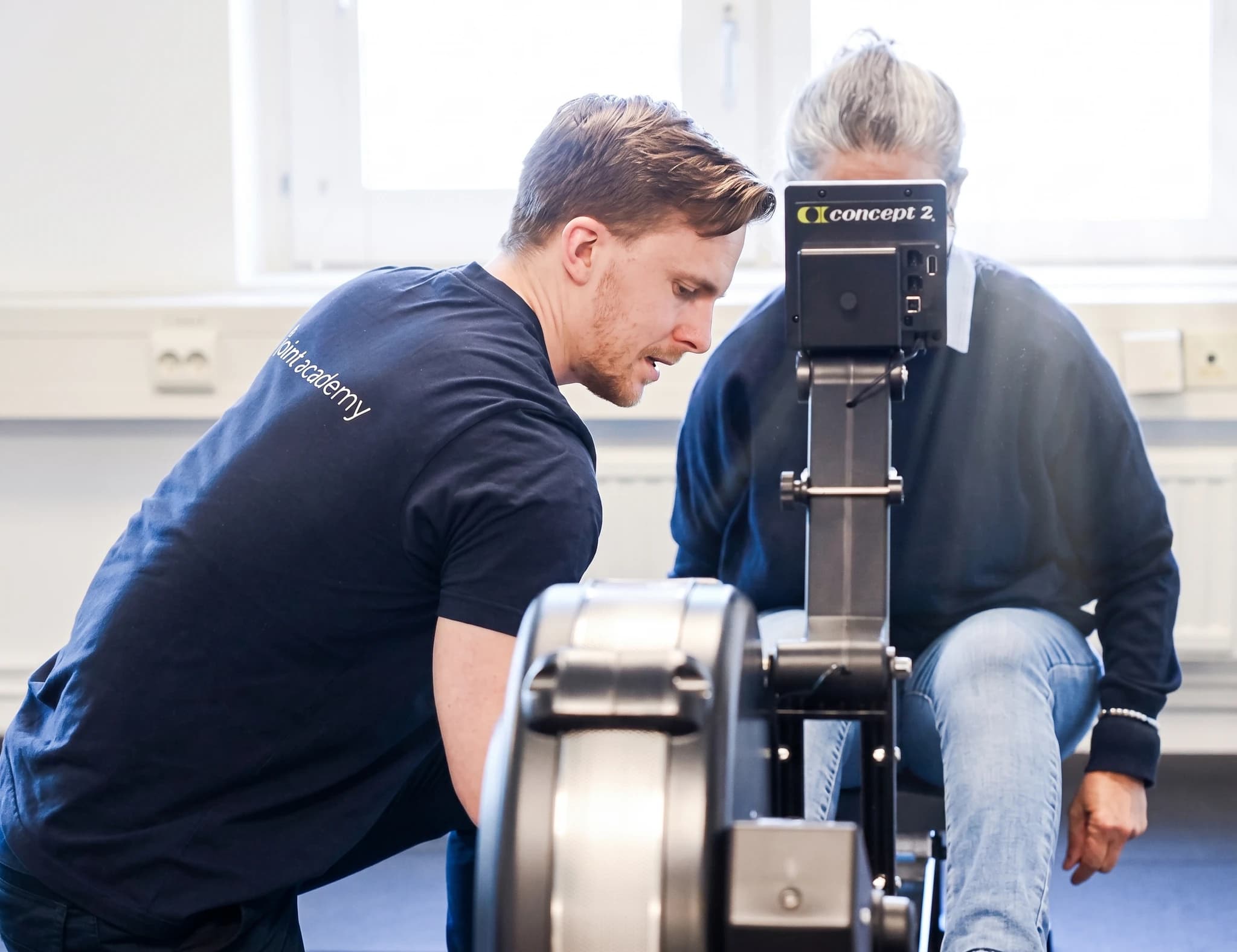 En tränare assisterar en patient på en roddmaskin i ett gym eller träningslokal.