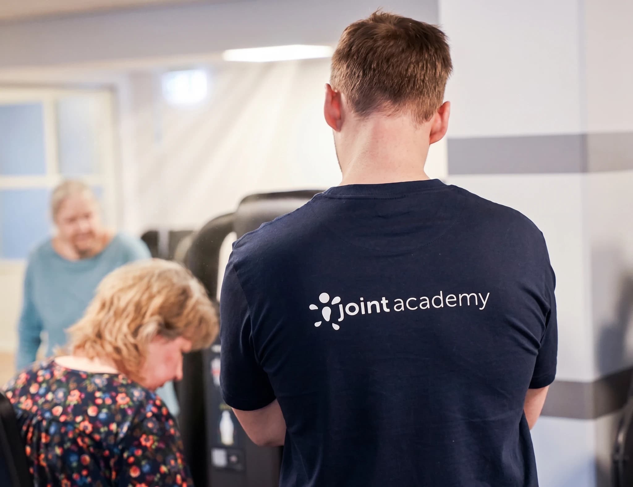 Fysioterapeut i mörkblå tröja med texten "joint academy" står bakom två kvinnliga patienter som tränar på gymutrustning.