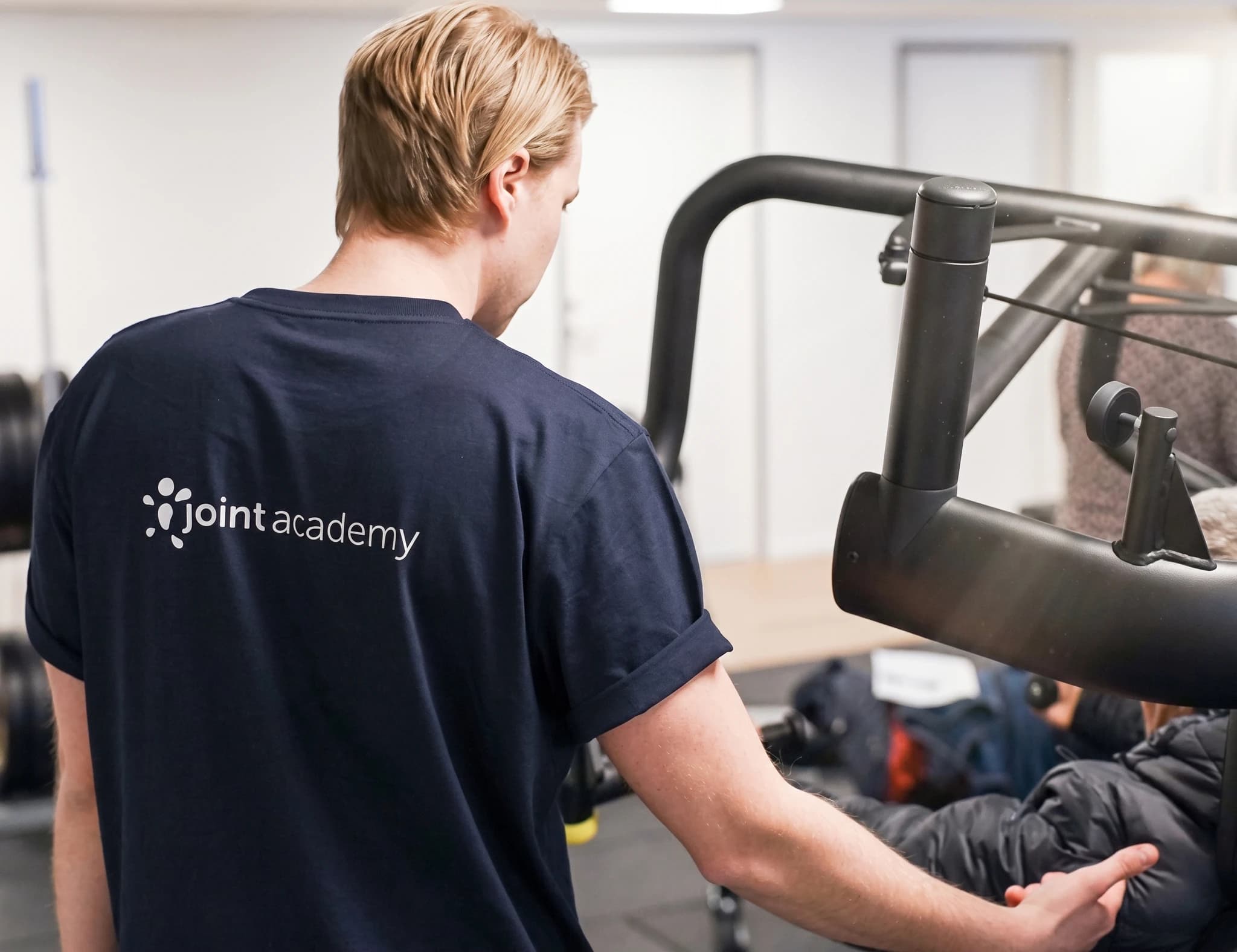 En ung man i en mörkblå t-shirt med "joint academy"-logotypen assisterar en patient på ett träningsredskap i en gymmiljö.