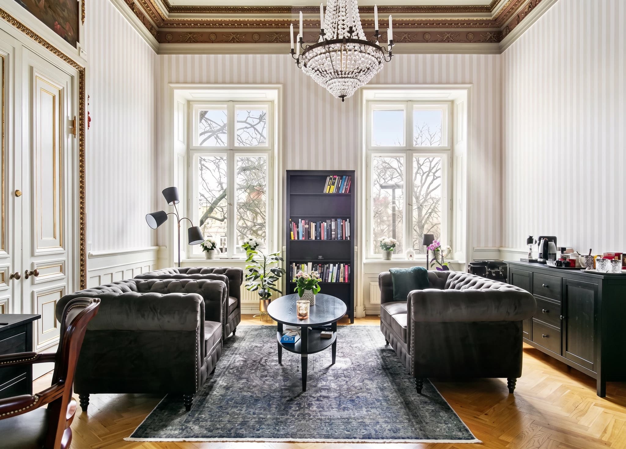 Väl upplyst vardagsrum med en stor kristallkrona i taket, randiga tapeter, två mörka soffor, ett runt soffbord, en golvlampa och en hög bokhylla fylld med böcker.