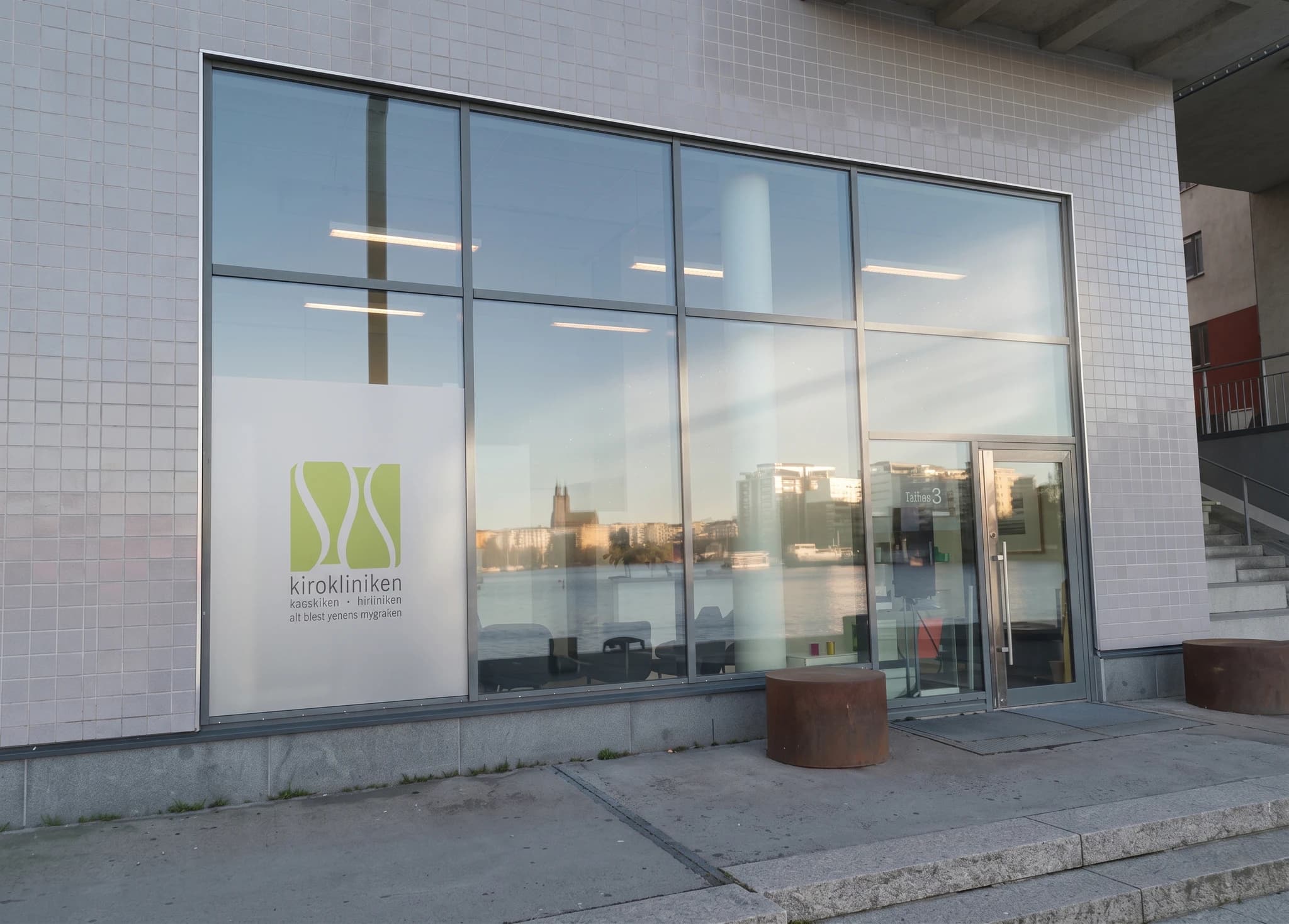 Exteriör av en klinikbyggnad med ett stort fönsterparti där en logotyp syns. Stadssiluetten och vattnet reflekteras i fönstren.