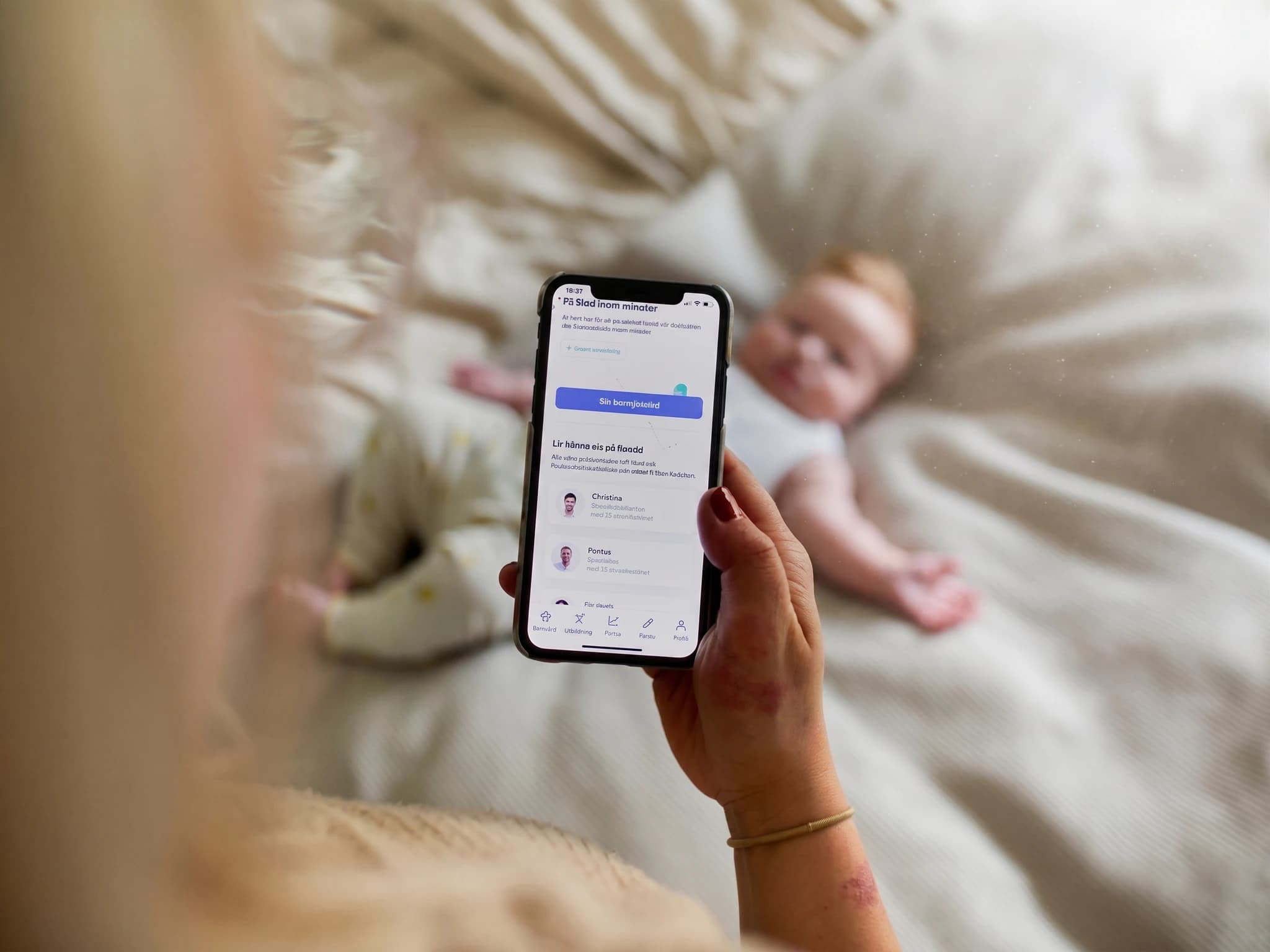 Kvinna som tittar på en mobiltelefon med en app för föräldrar, med en bebis liggande i bakgrunden.