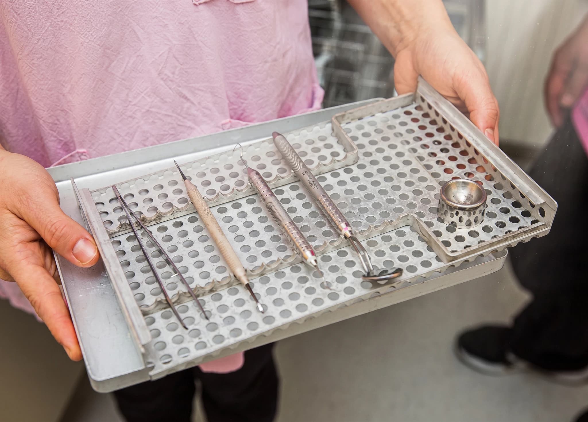 En person håller i en bricka med tandläkarinstrument, inklusive pincett och sonder, i en steriliseringskorg.