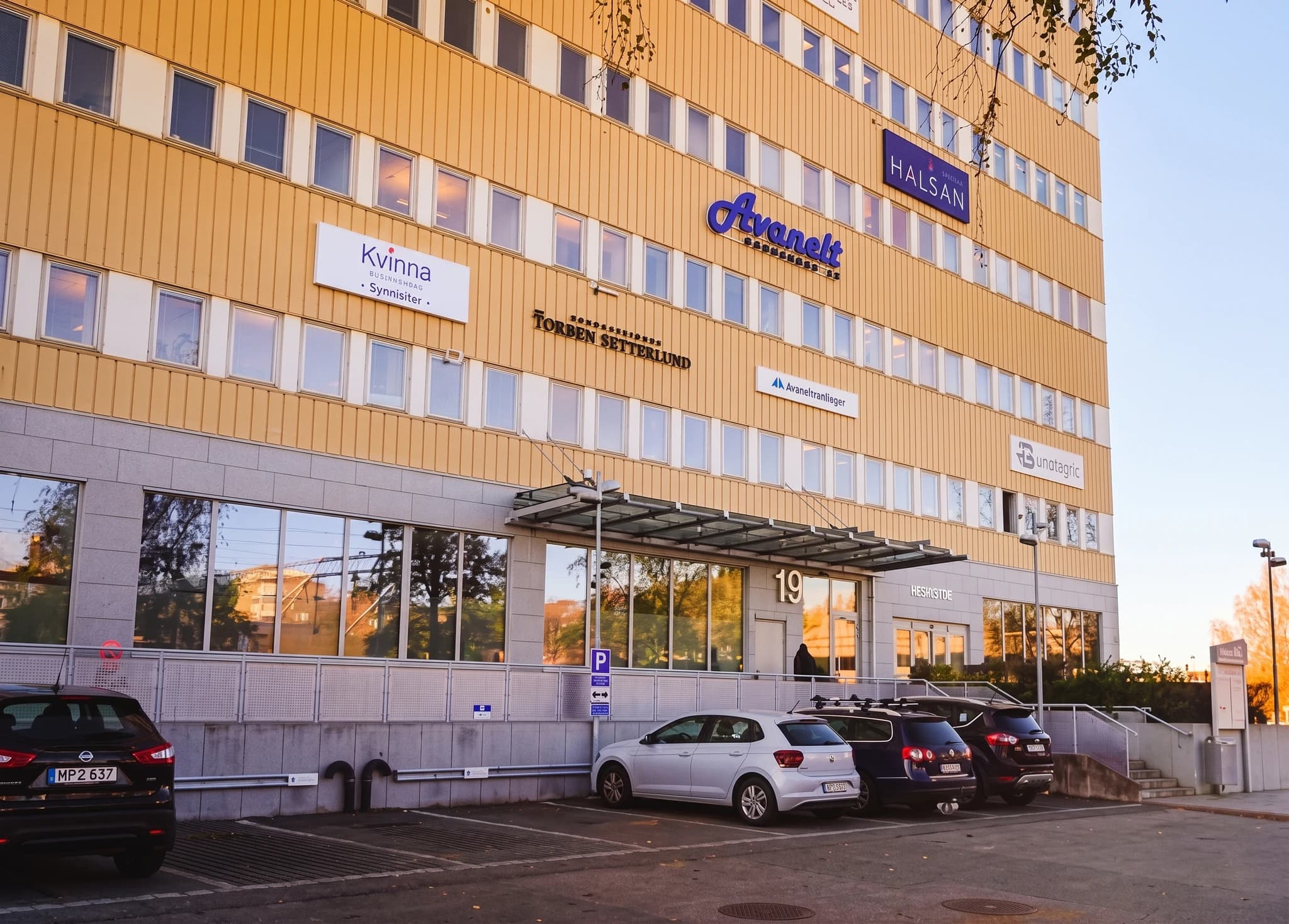 Exteriör av en gul byggnad med många fönster och skyltar för olika företag, inklusive 'Avanett' och 'Halsan'. Parkerade bilar syns framför entrén.