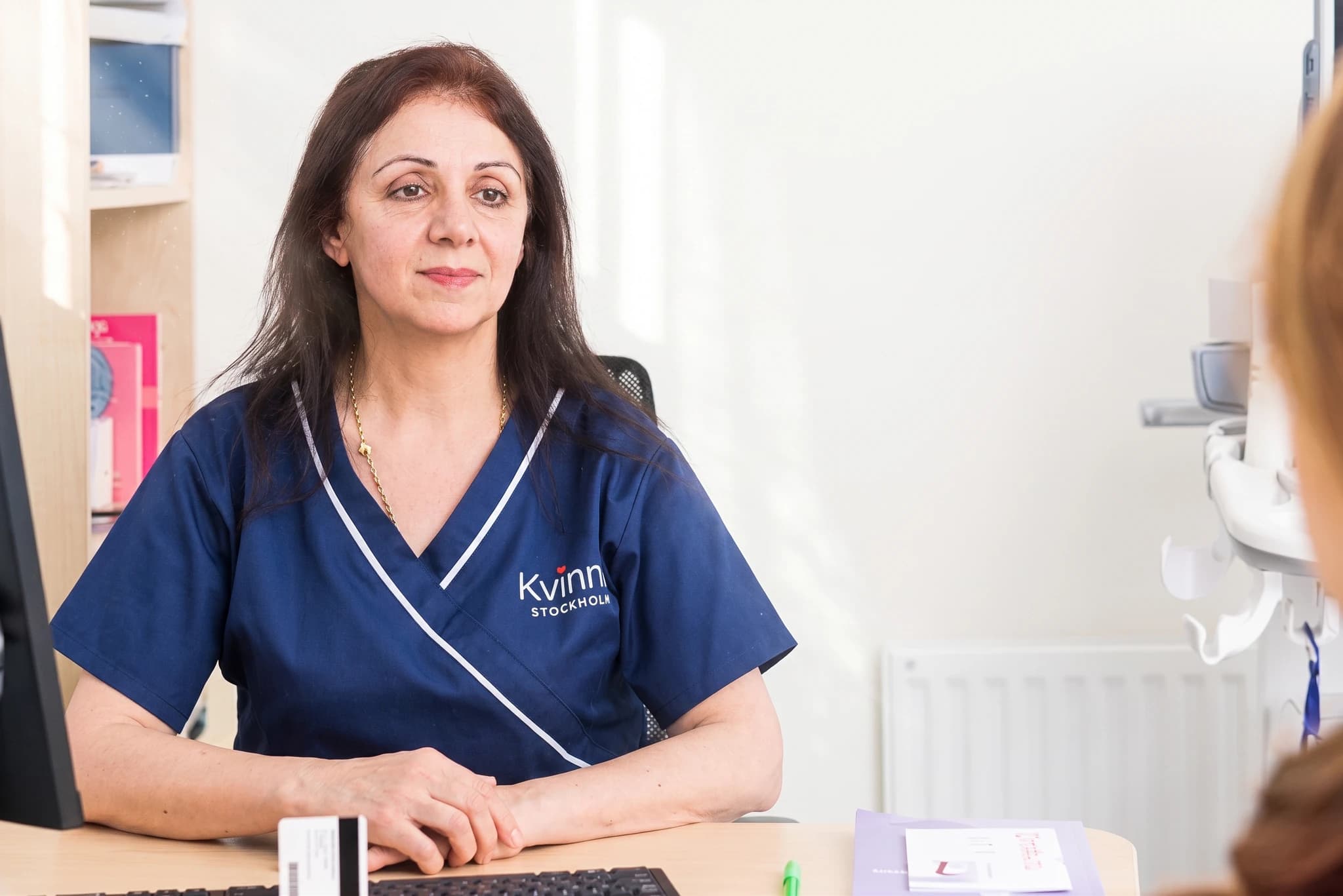 Kvinnlig läkare i blå kläder sitter vid ett skrivbord och pratar med en patient vars ansikte inte syns.