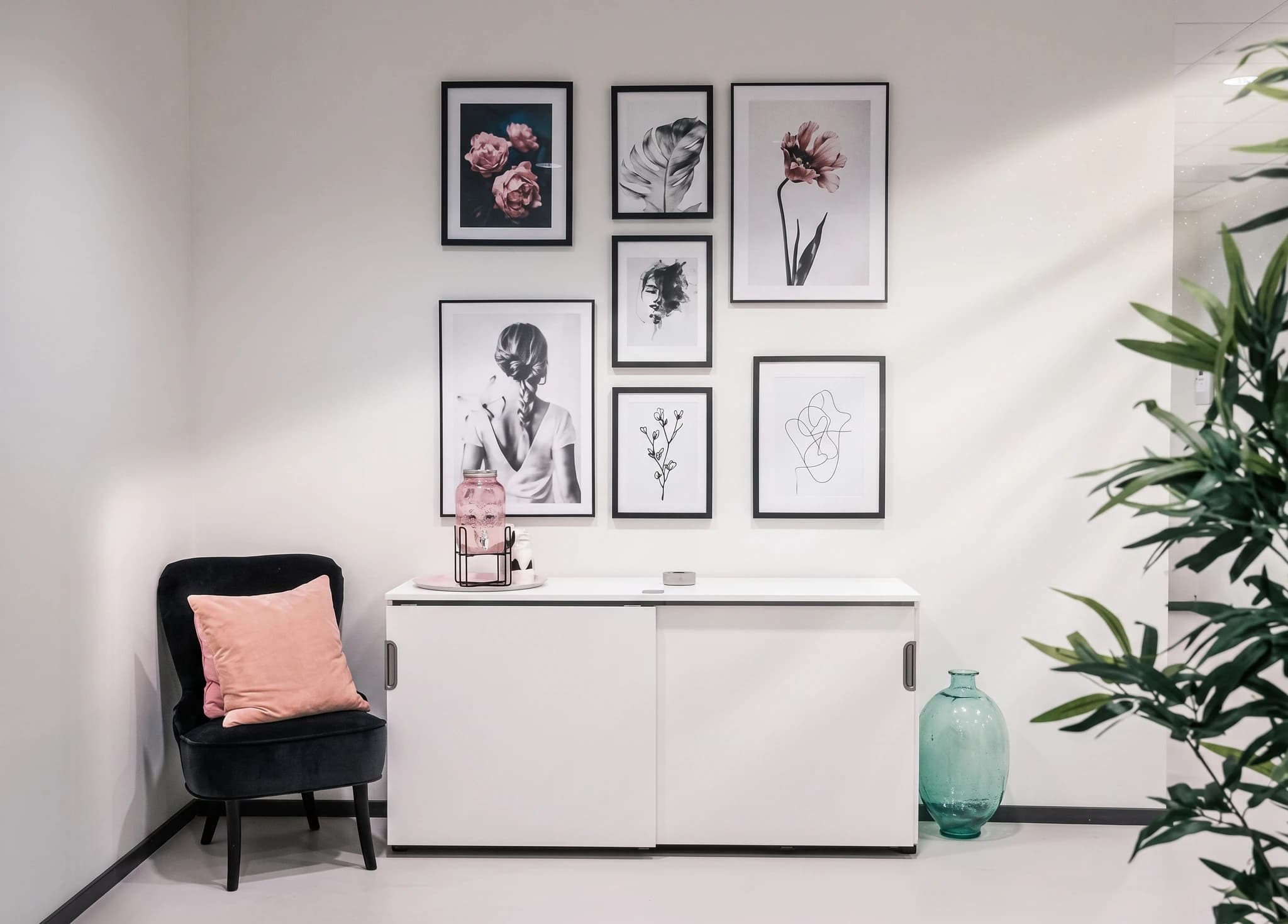 Vitt förvaringsskåp med konstverk på väggen, en svart stol med en rosa kudde och en grön glasvas.