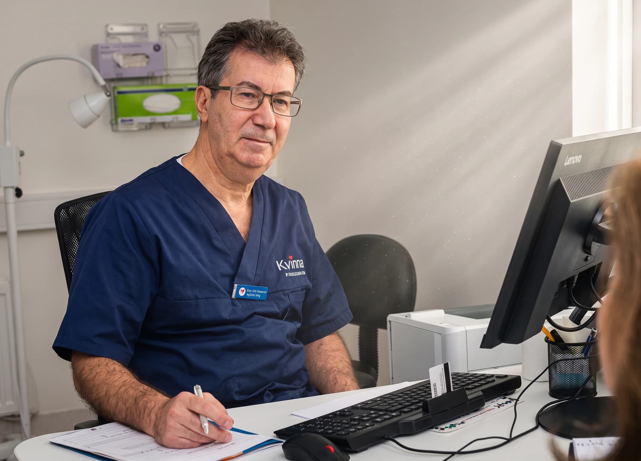 Manlig läkare i blå uniform sitter vid ett skrivbord och pratar med en patient som syns delvis.