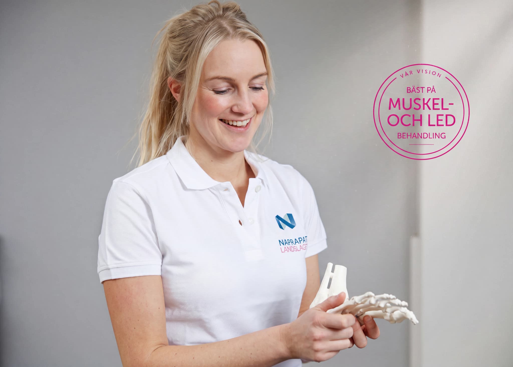 En ung kvinna med blont hår, klädd i en vit pikétröja med logotypen för Naprapat Landslaget, undersöker en modell av en fot. Hon ler och ser glad ut.