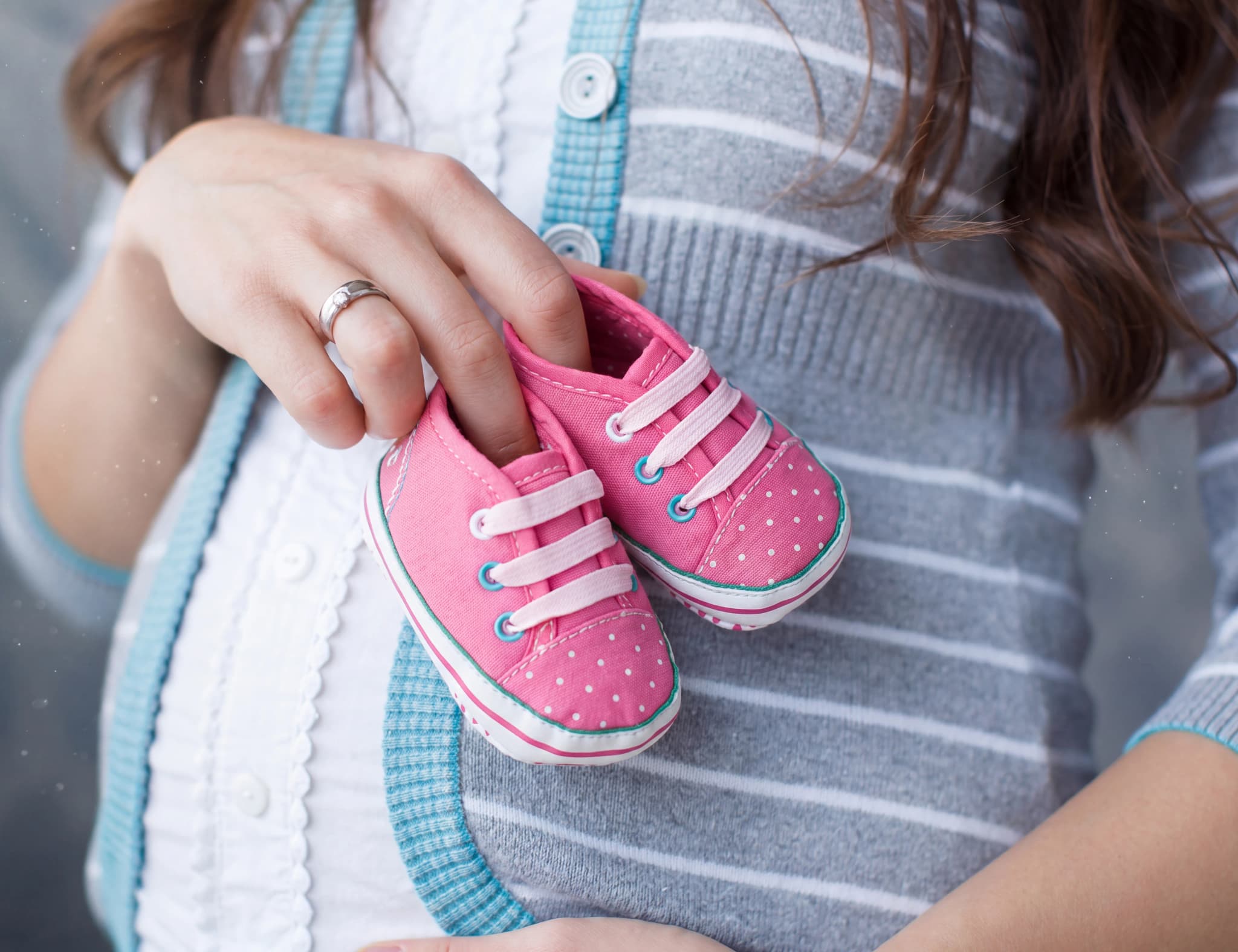 En gravid kvinna håller två små rosa babyskor med prickar och vita snören framför magen.