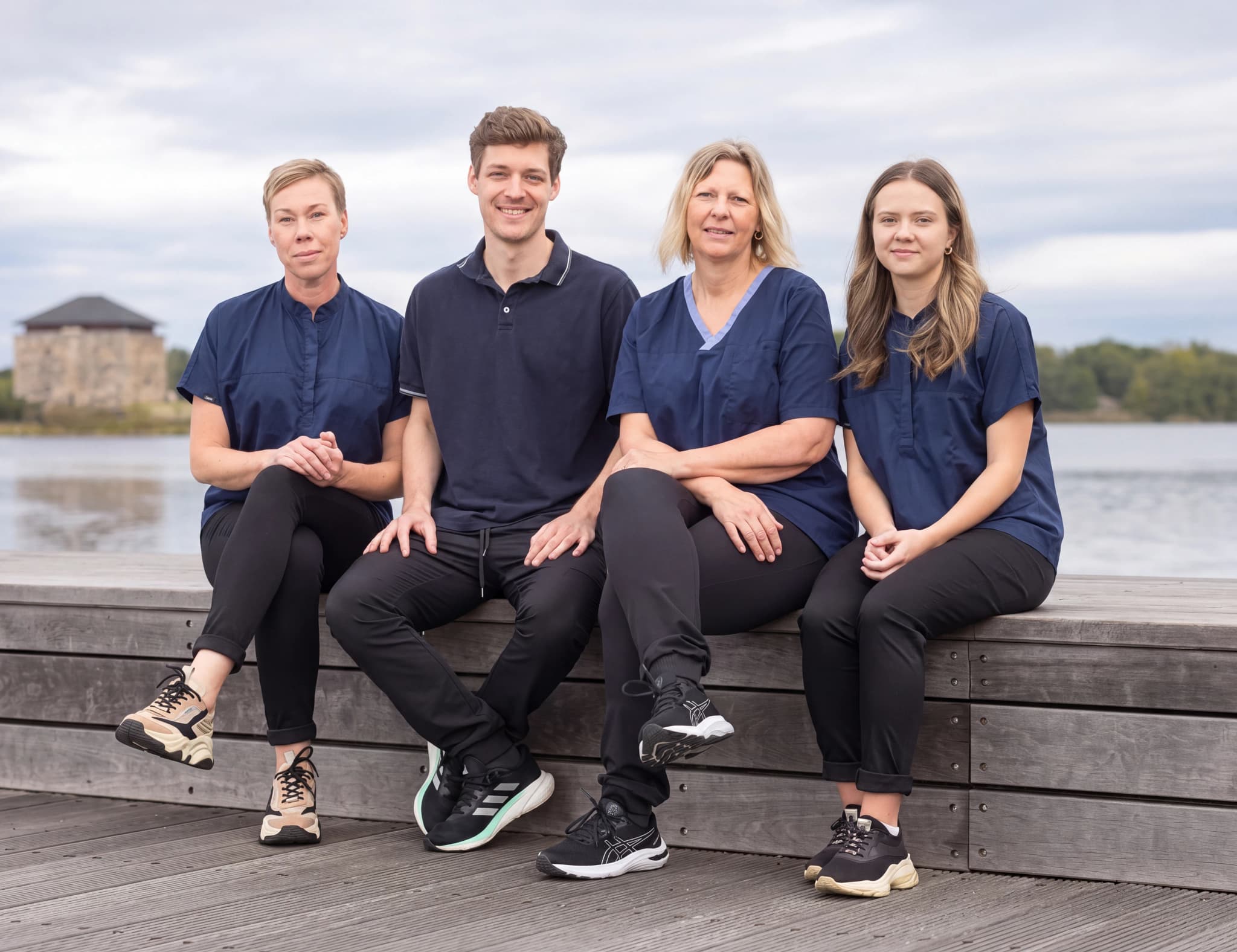 Fyra vårdpersonal sitter på en träbänk utomhus med en byggnad och vatten i bakgrunden.