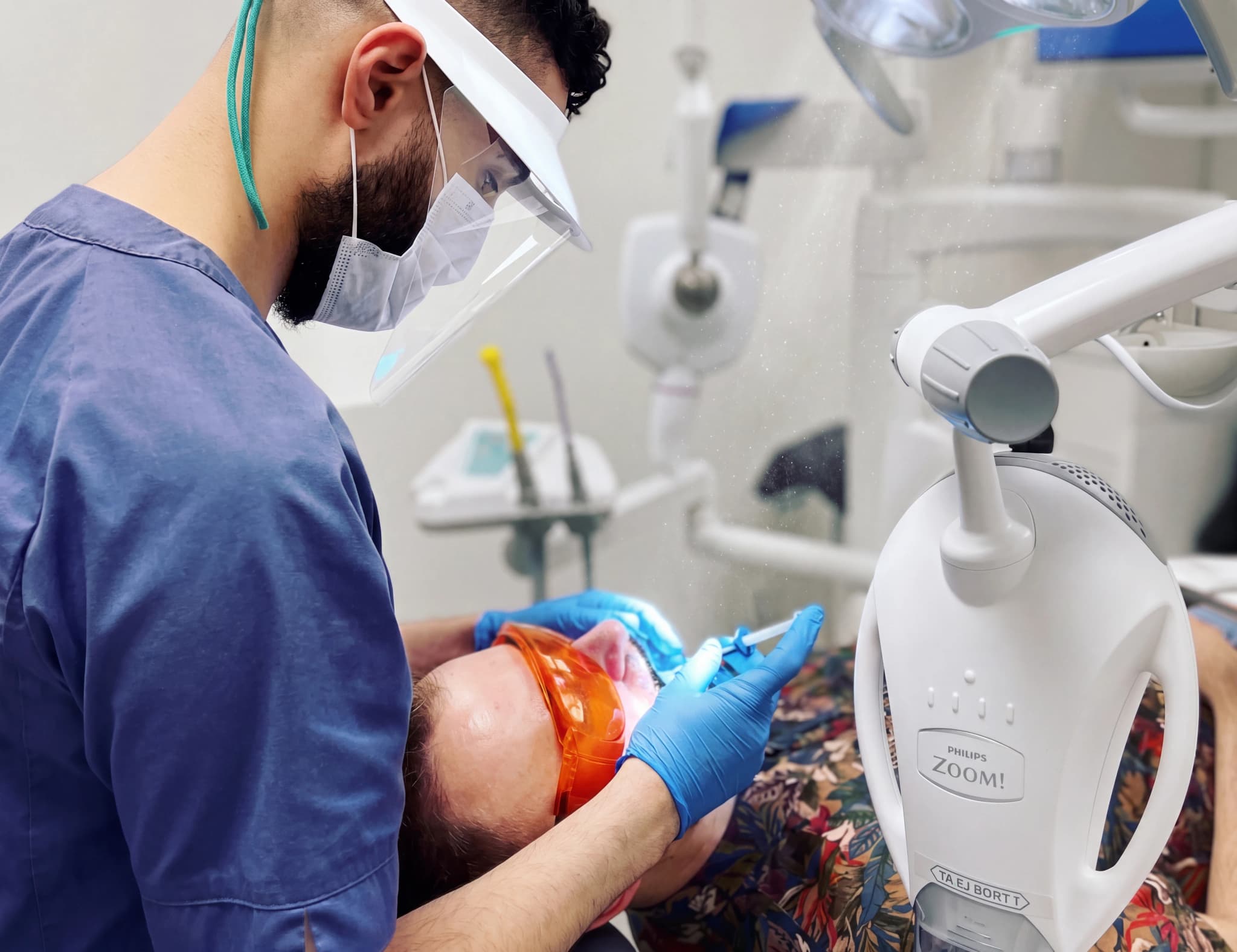 Tandläkare med skyddsutrustning utför en tandblekningsprocedur på en patient med skyddsglasögon.