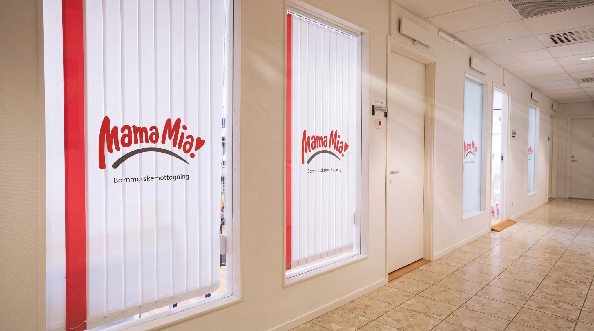Mama Mia-logotypen med texten Barnmorskemottagning på vita vertikala persienner i en korridor.
