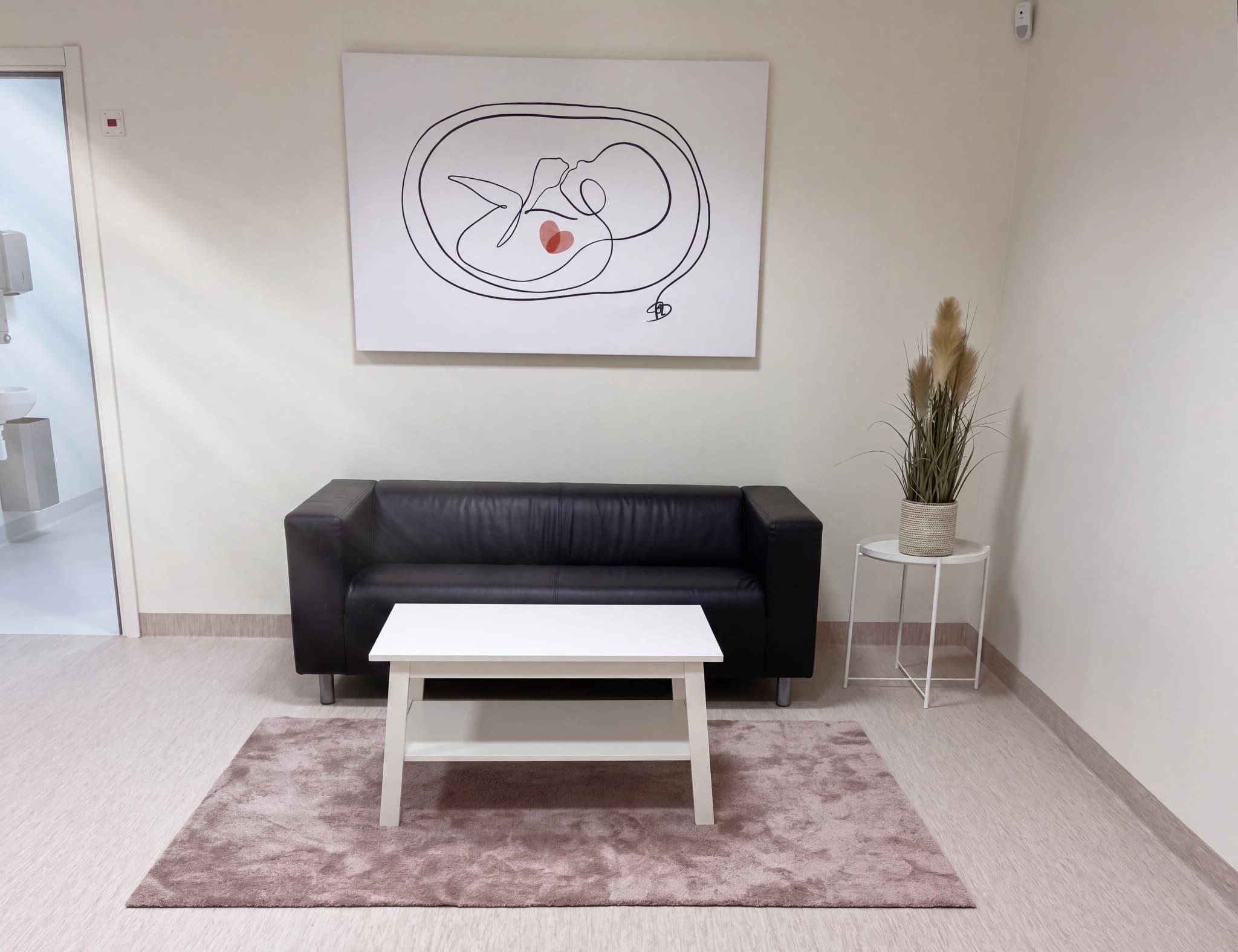Ett väntrum med en svart soffa, ett vitt soffbord, en matta och konstverk på väggen som avbildar en bebis i livmodern.