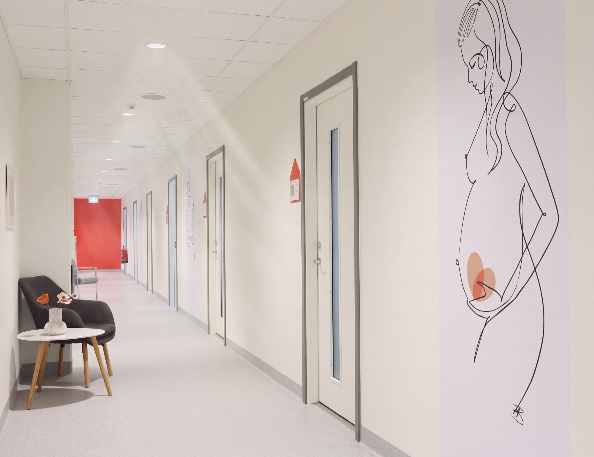 En modern korridor med väggkonst av en gravid kvinna och en väntrumsdel med stol och bord.