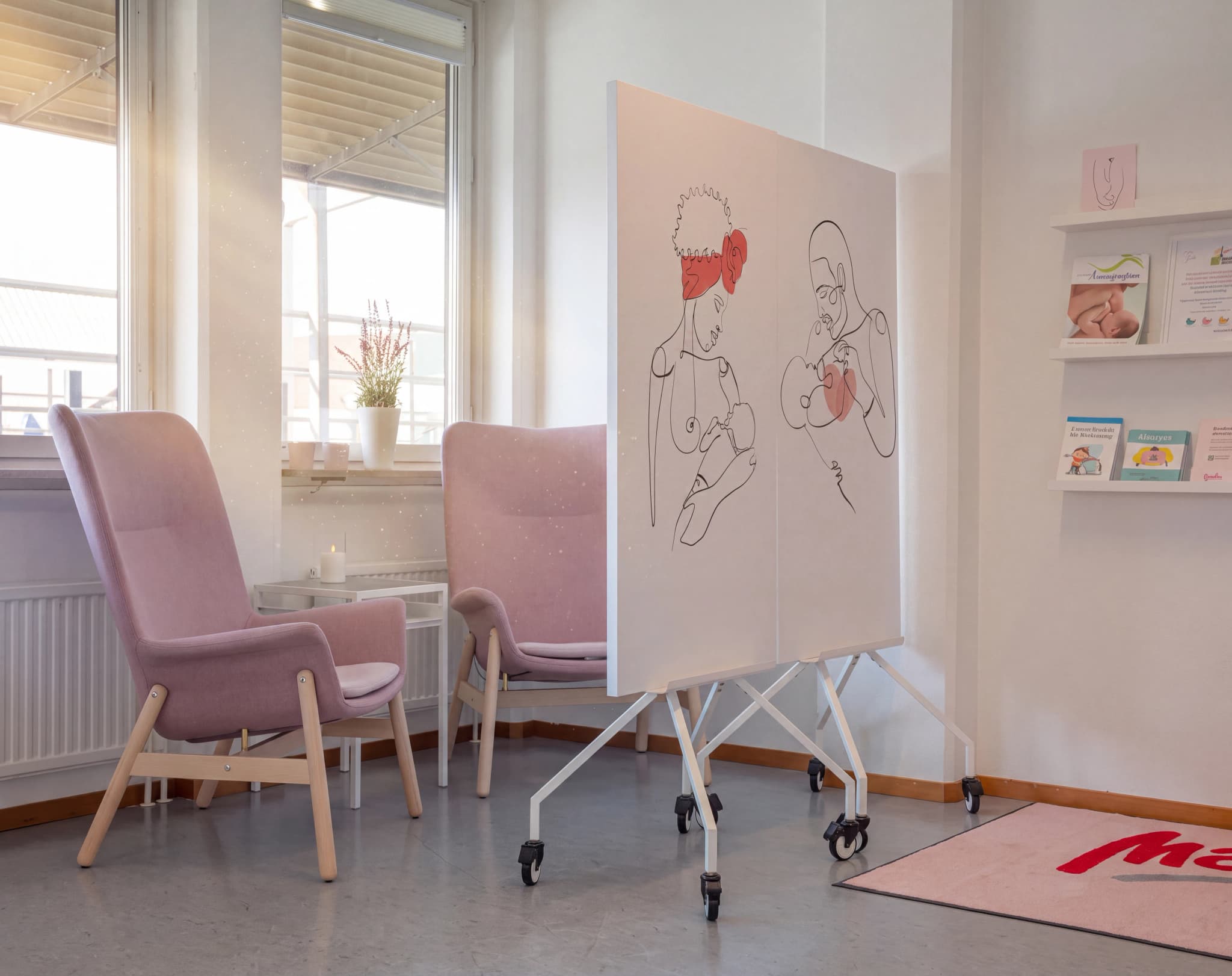 Två ljusrosa fåtöljer med träben i ett väntrum, med ett fönster med persienner och en krukväxt i bakgrunden.