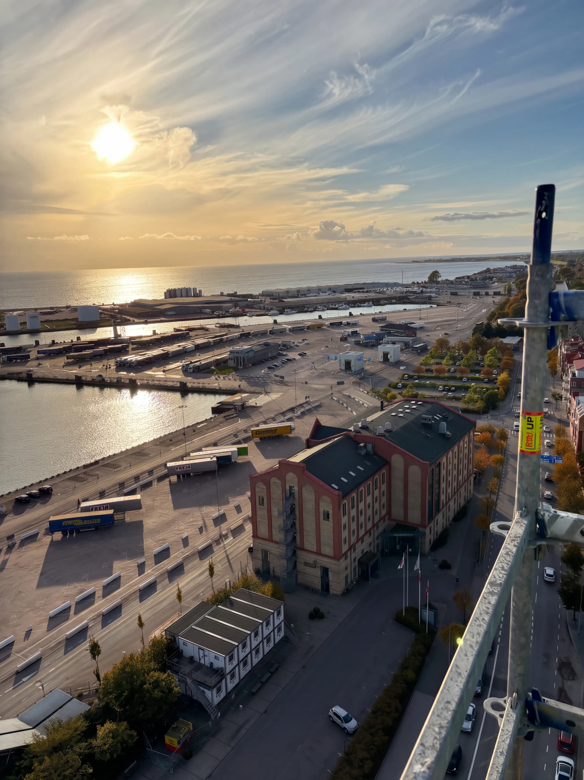 Industriell hamnmiljö vid solnedgången med en stor byggnad, många lastbilar och havet.