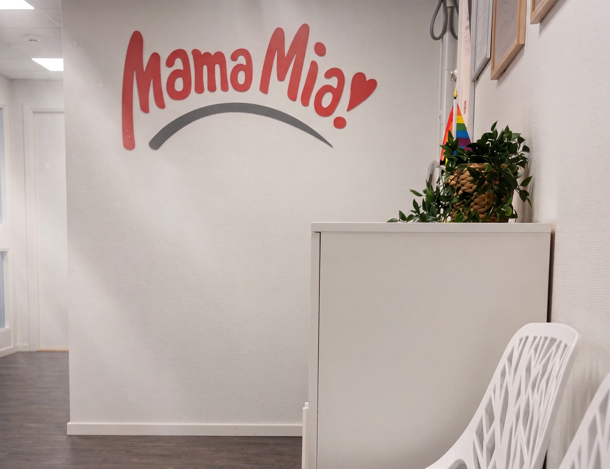 Texten 'Mama Mia' med ett hjärta ovanför på en vit vägg, med en grå båge under. I bakgrunden syns en vit möbel och en stol.