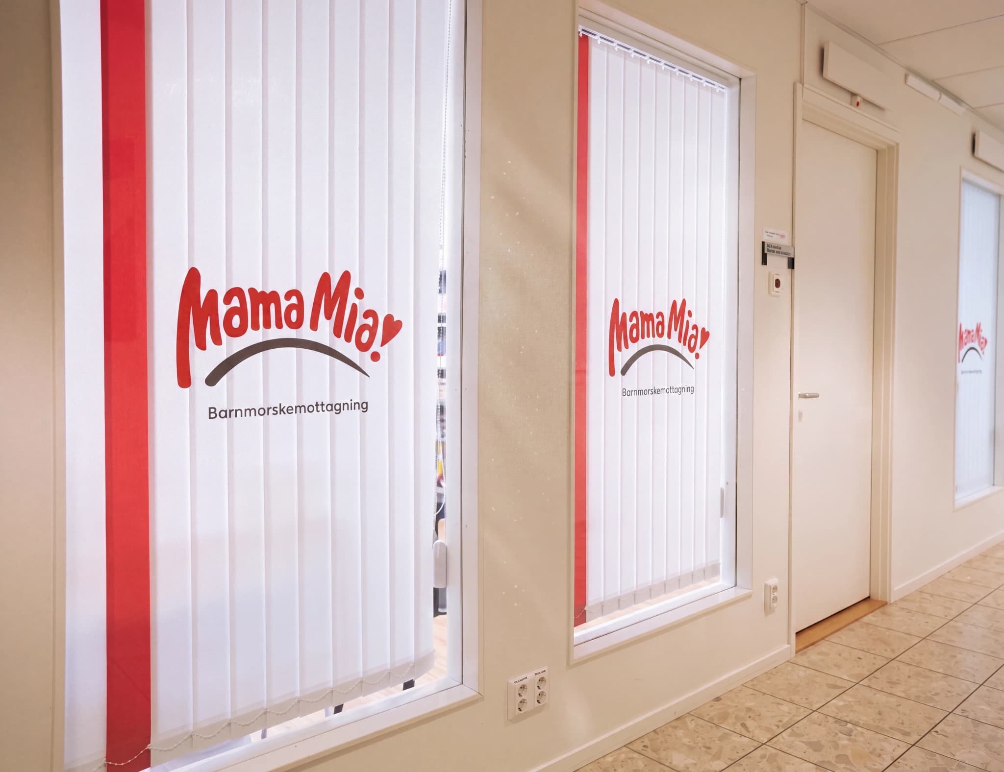 Fönster med vertikala persienner för Mama Mia barnmorskemottagning med en röd rand och logotyp.